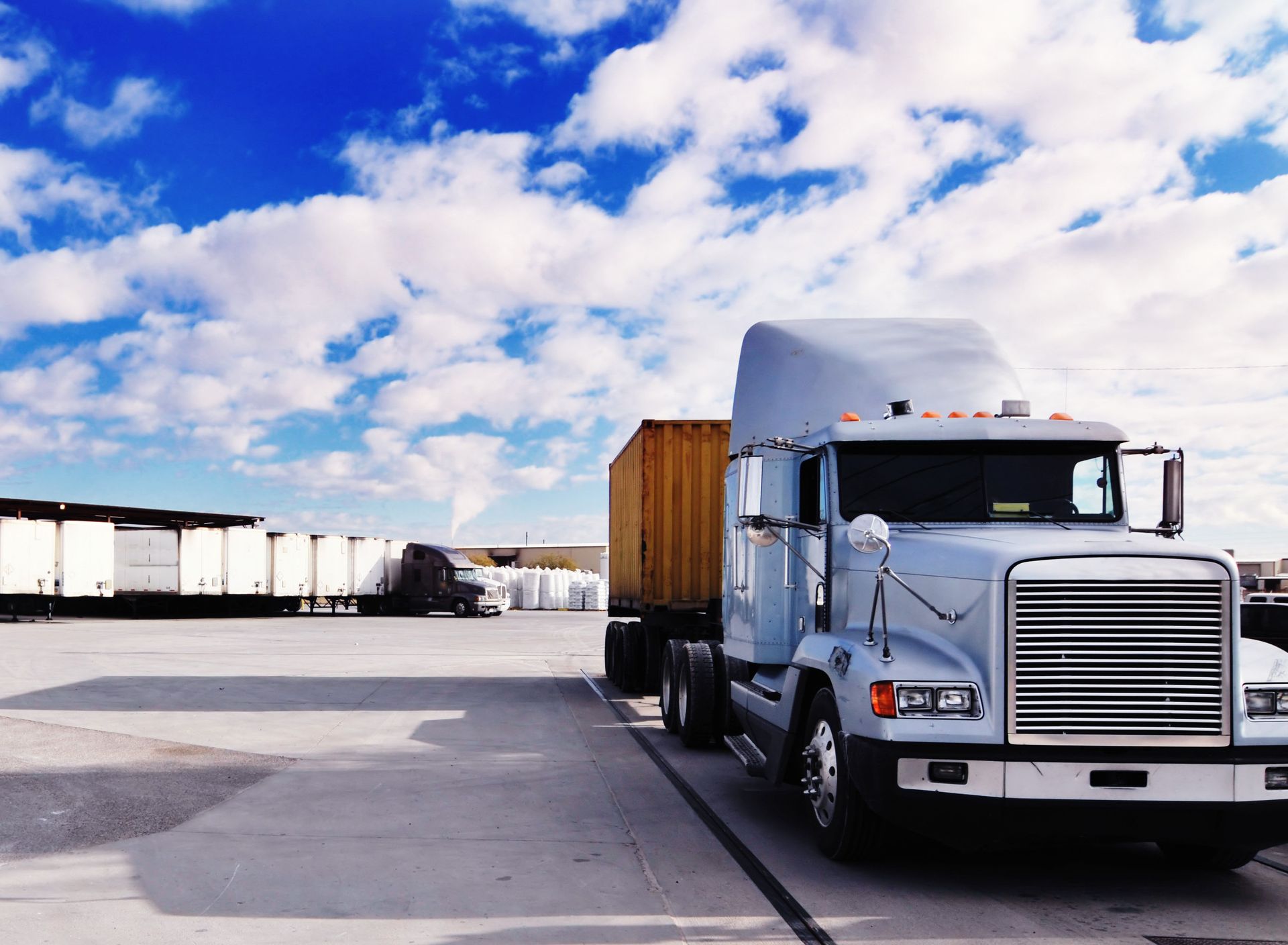 Semi-truck with cargo trailer parked at a loading dock against a cloudy blue sky.