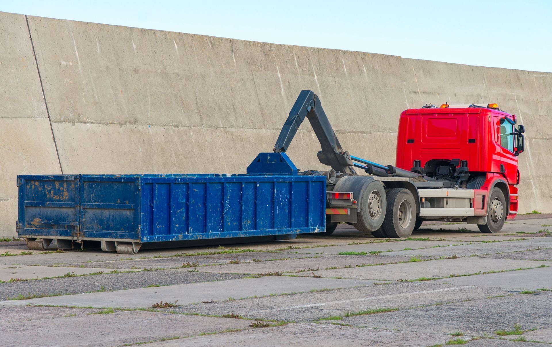 Red truck with a hydraulic arm lifting a blue dumpster in front of a concrete wall.