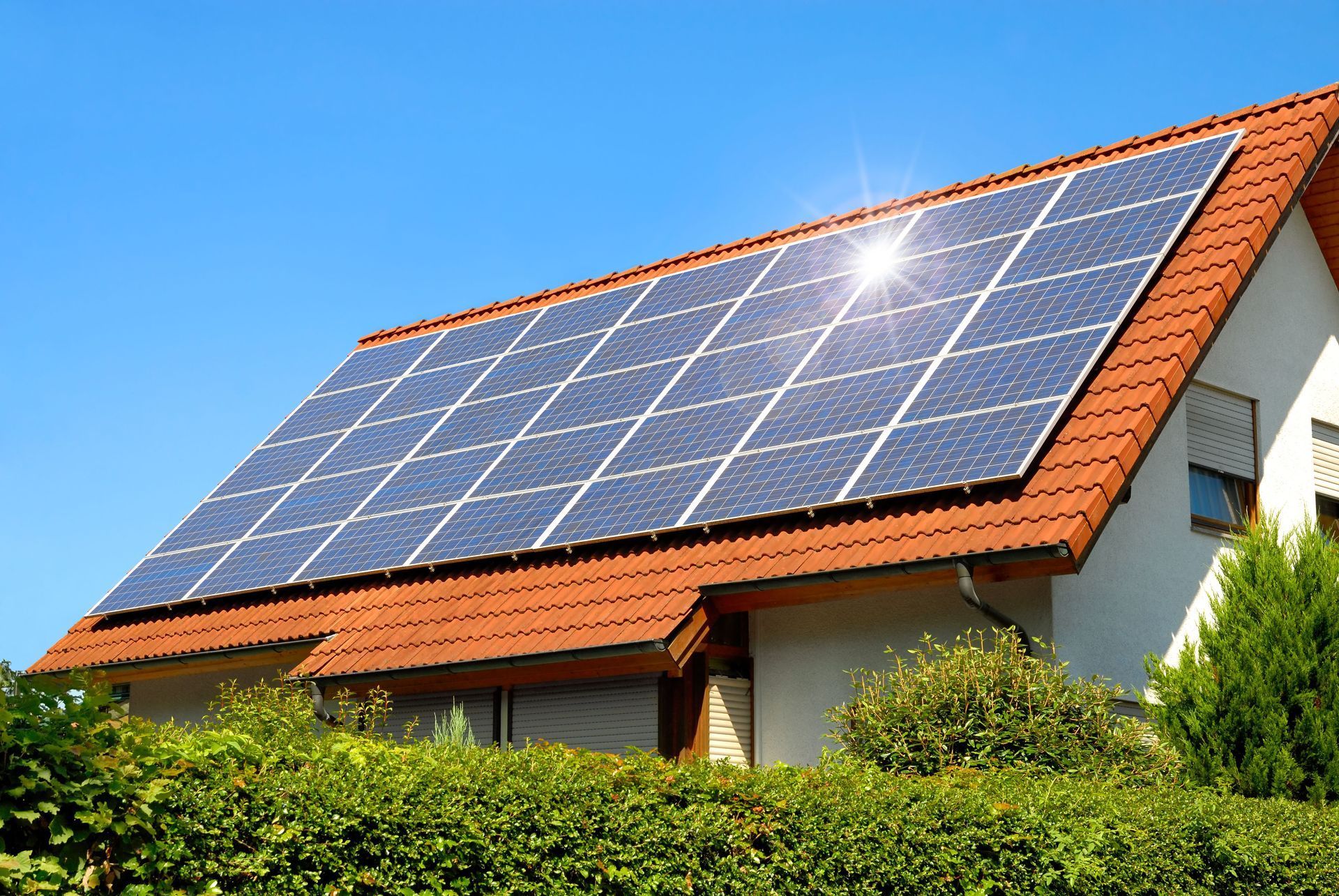 Solar panels on a red-tiled roof of a house, bright sun against a clear blue sky.
