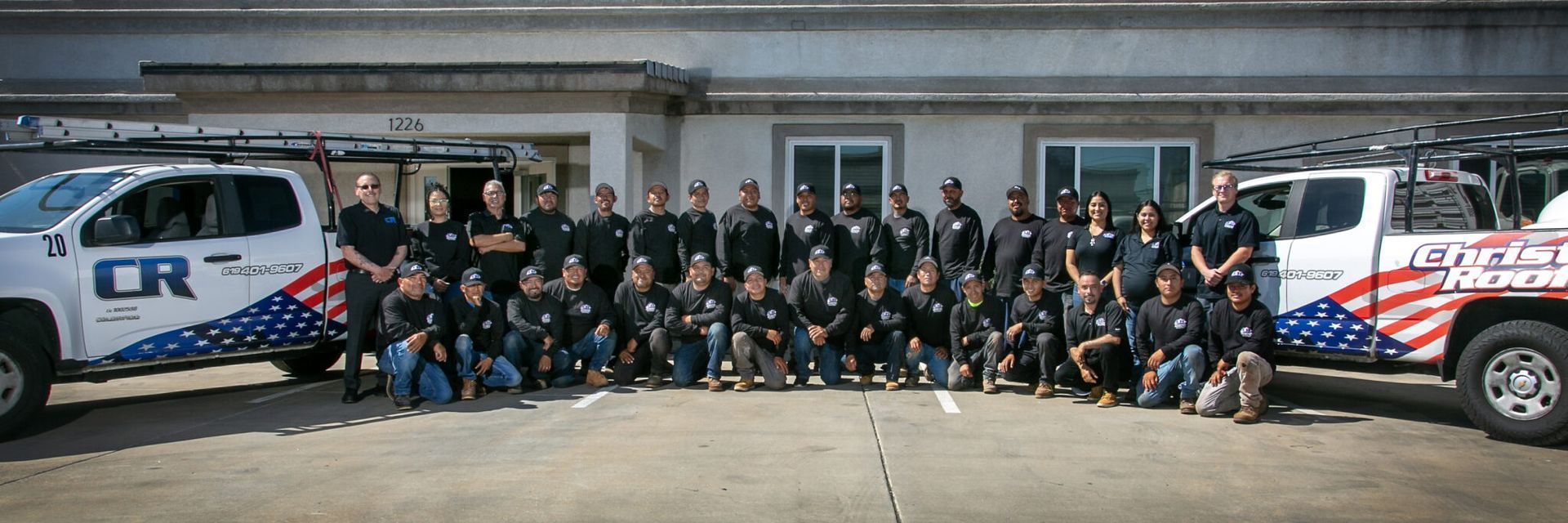 Group of people in black shirts and blue jeans, standing in front of building and trucks.