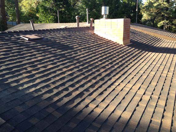 Brown asphalt shingle roof with a brick chimney and metal vent against a backdrop of trees.