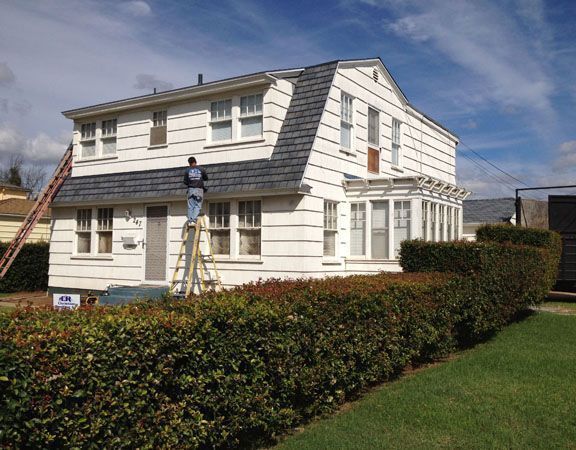 Person on a ladder working on the roof of a two-story white house with a dark gray roof.