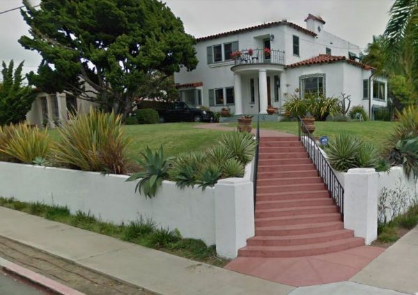 White stucco house with red-tiled stairs, a green lawn, and a low wall with plants.