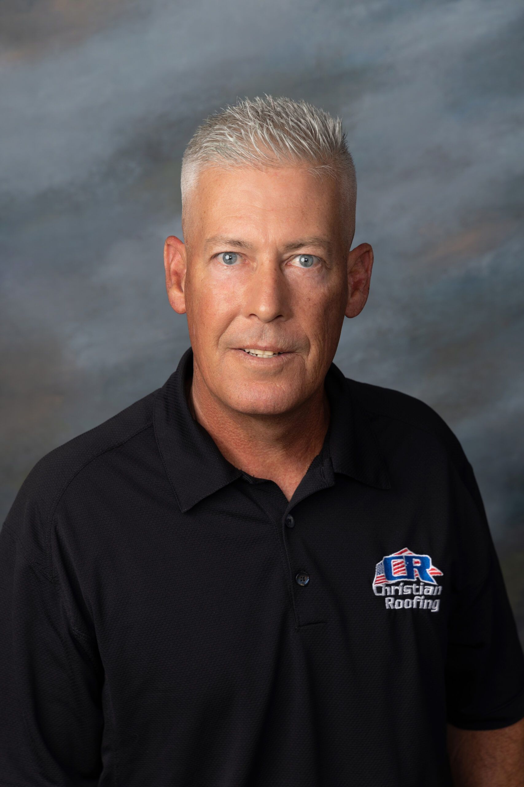 Man with gray hair wearing a black polo shirt, smiling, in front of a blue and gray background.