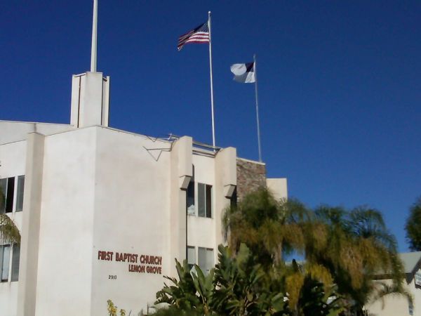 First Baptist Church building with US and Christian flags against a blue sky.