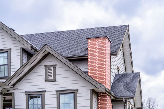 House exterior with gray siding, brick chimney, and gray shingle roof against a cloudy sky.