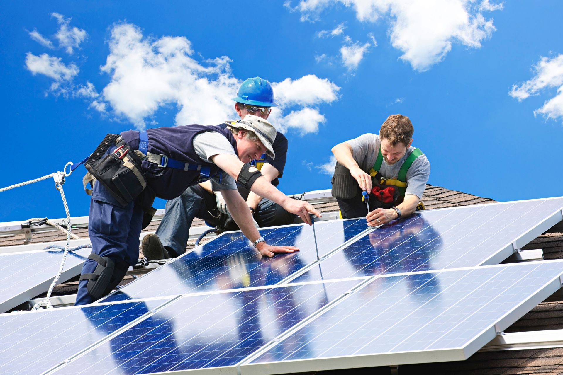 Workers installing solar panels on a roof against a bright blue sky.