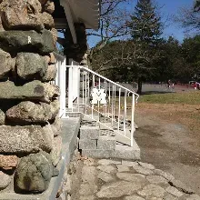A stone-walled building exterior featuring a white metal railing leading to steps, set against a sunny, outdoor park area.
