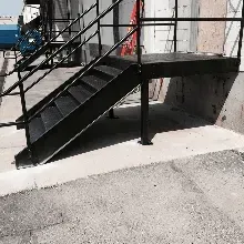 Black metal stairs with handrails leading to an elevated platform against a building wall on a concrete surface.
