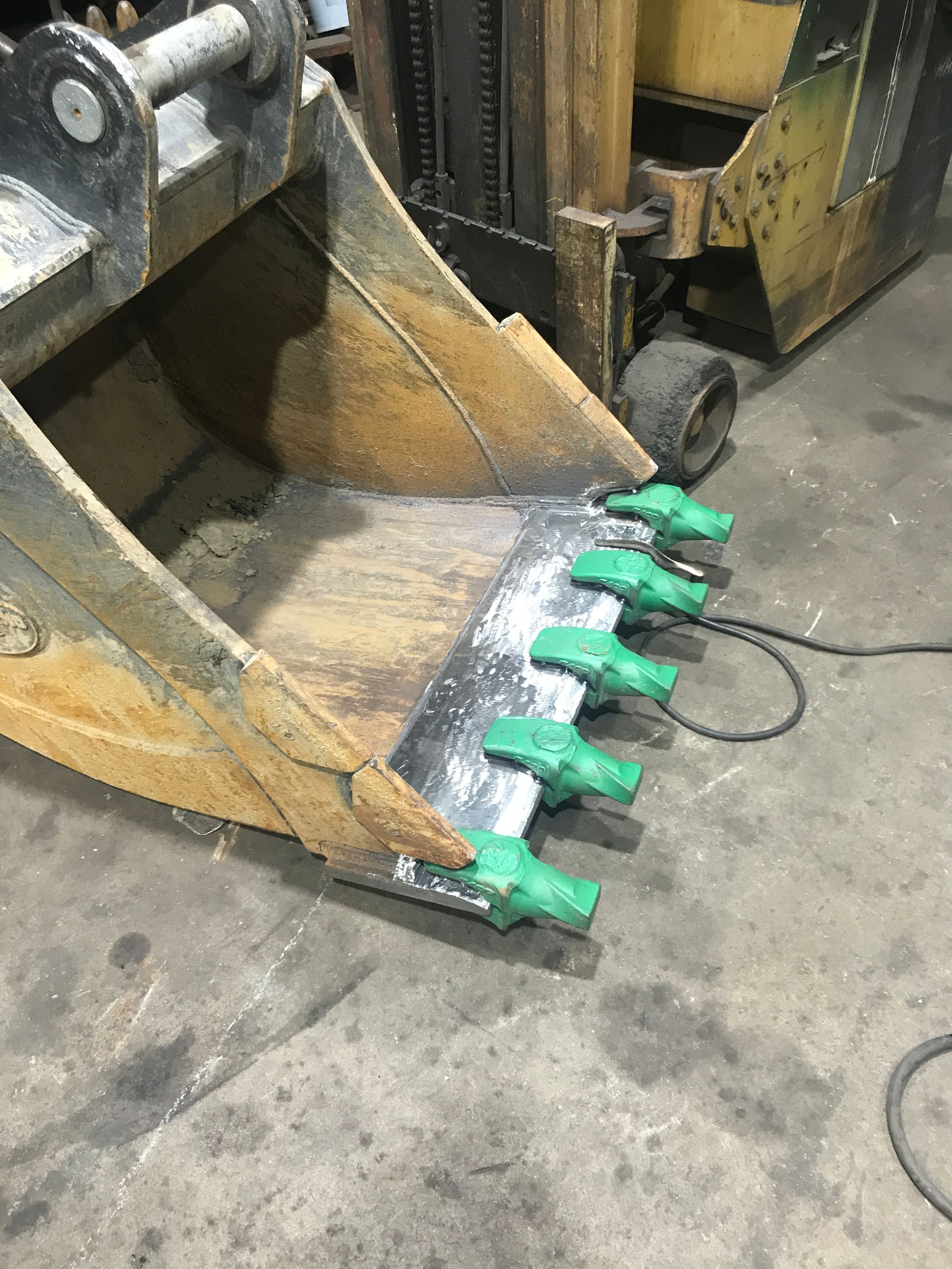 An excavator bucket with five bright green teeth installed on the cutting edge, positioned on a concrete shop floor.