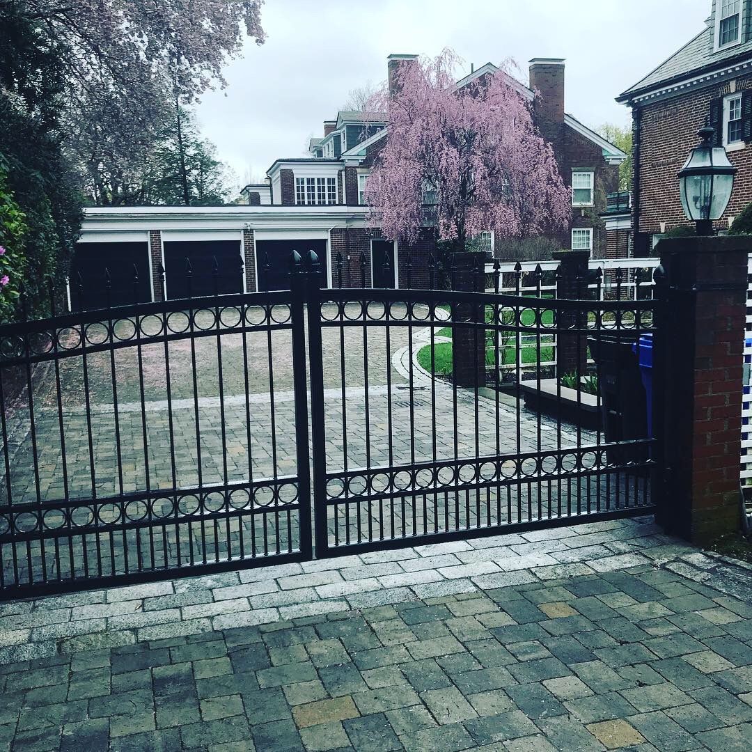 A black wrought-iron gate stands before a driveway, leading to a brick house with a prominent, blooming pink tree.