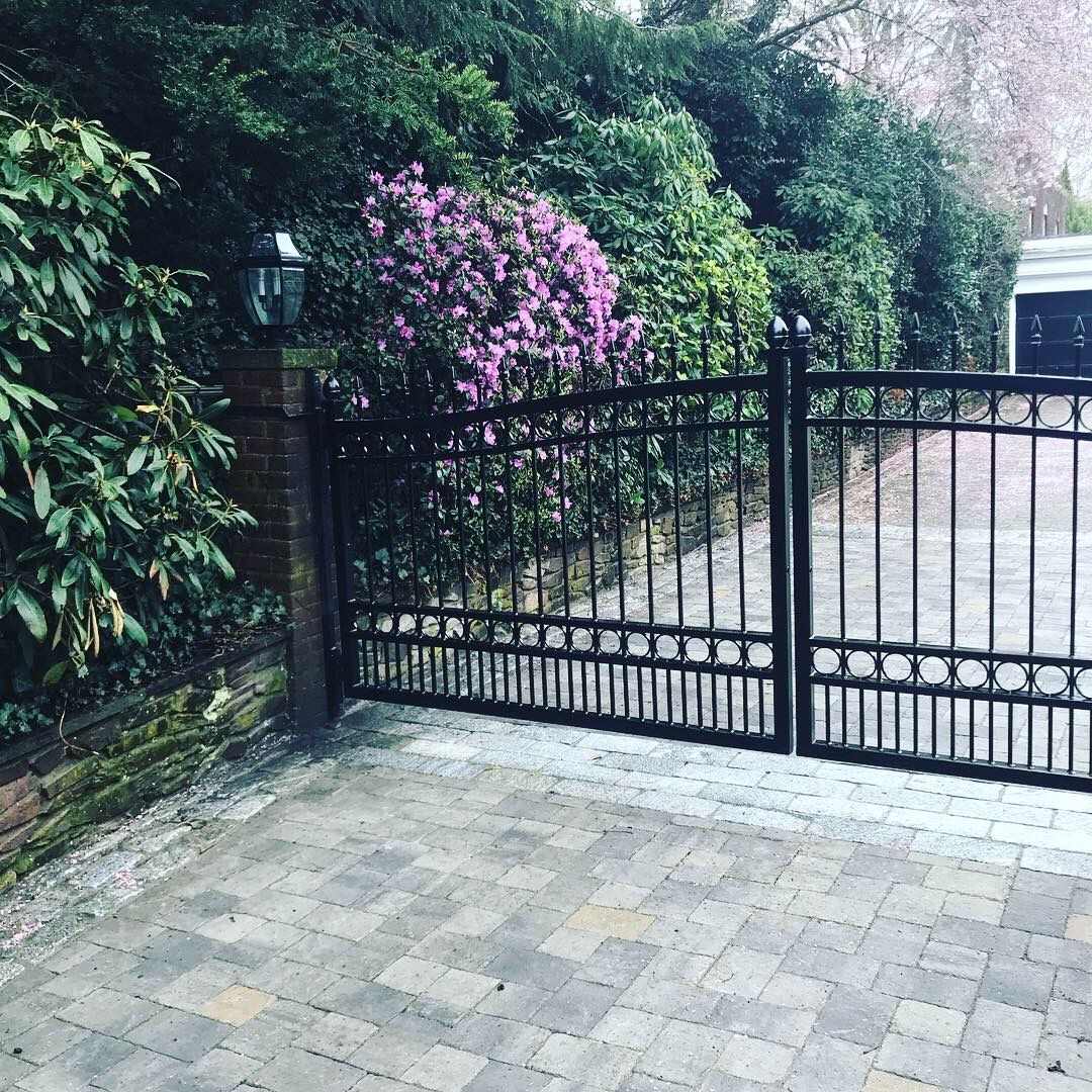 Black metal double gates open onto a stone-paved driveway, flanked by a brick post, lush green hedges, and purple flowers.