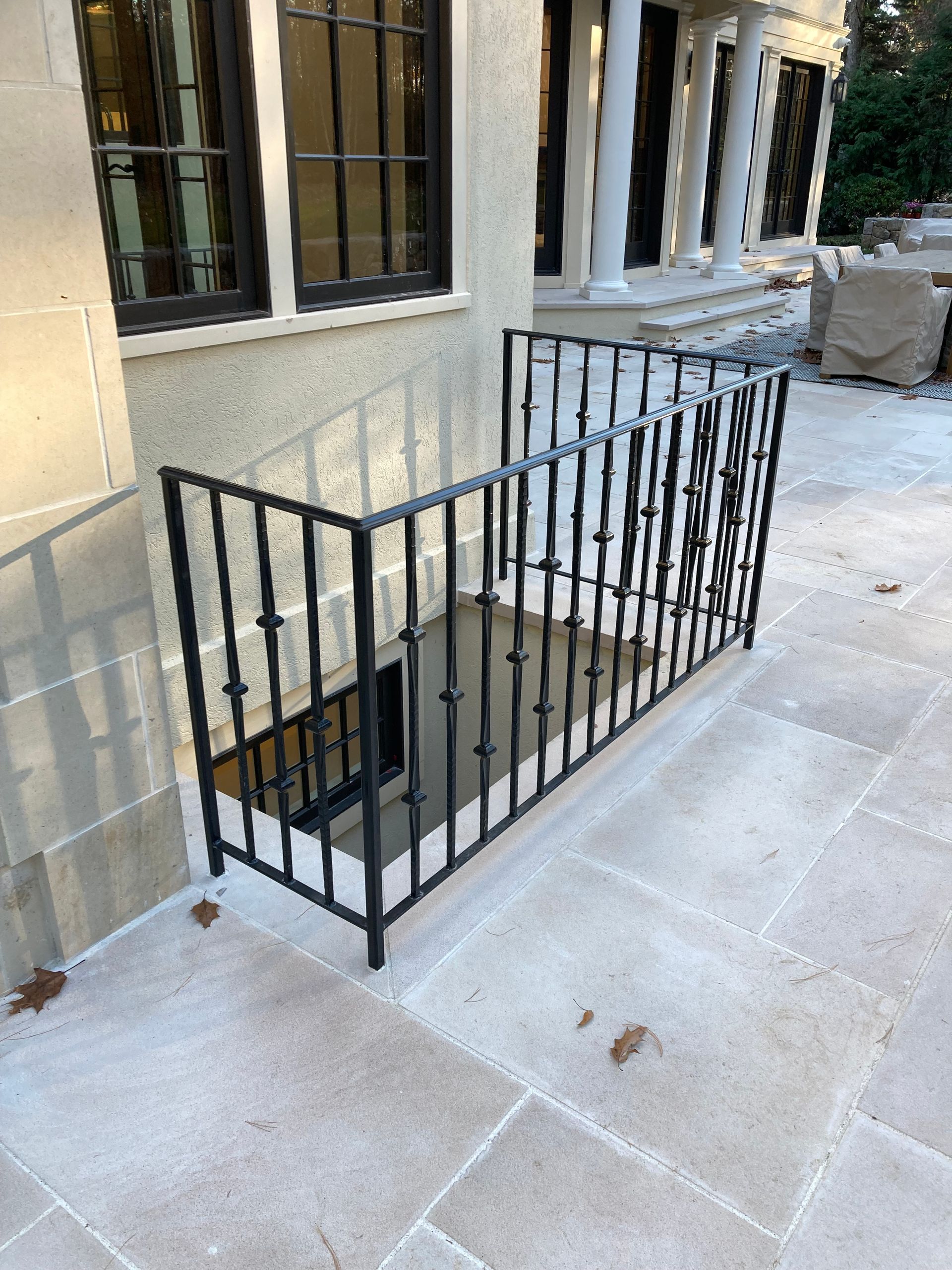 A black metal railing surrounds a basement window well on a stone patio outside a house.