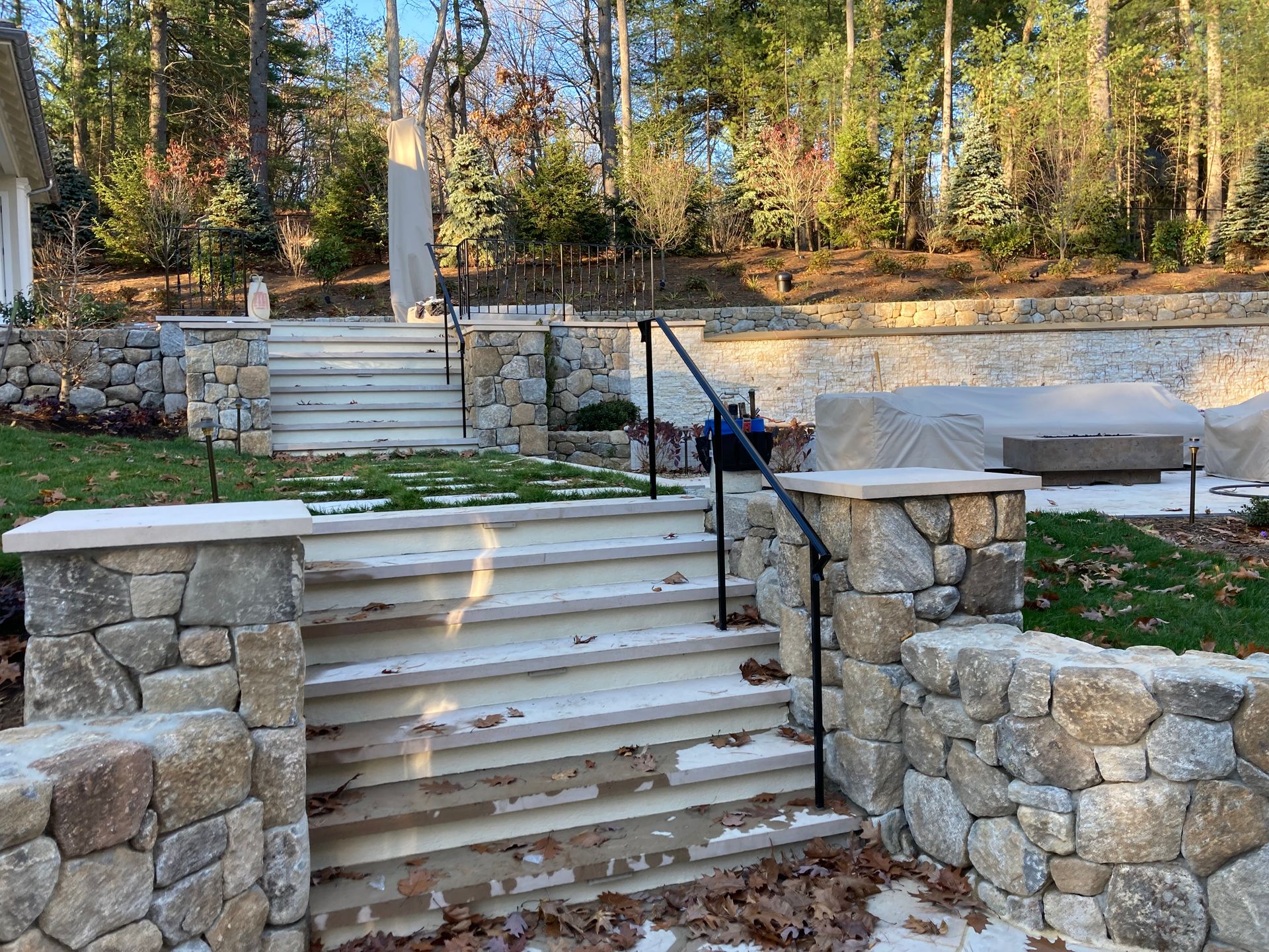 Stone steps with a black metal railing ascend through a landscape of tiered rock walls and grass in a wooded setting.