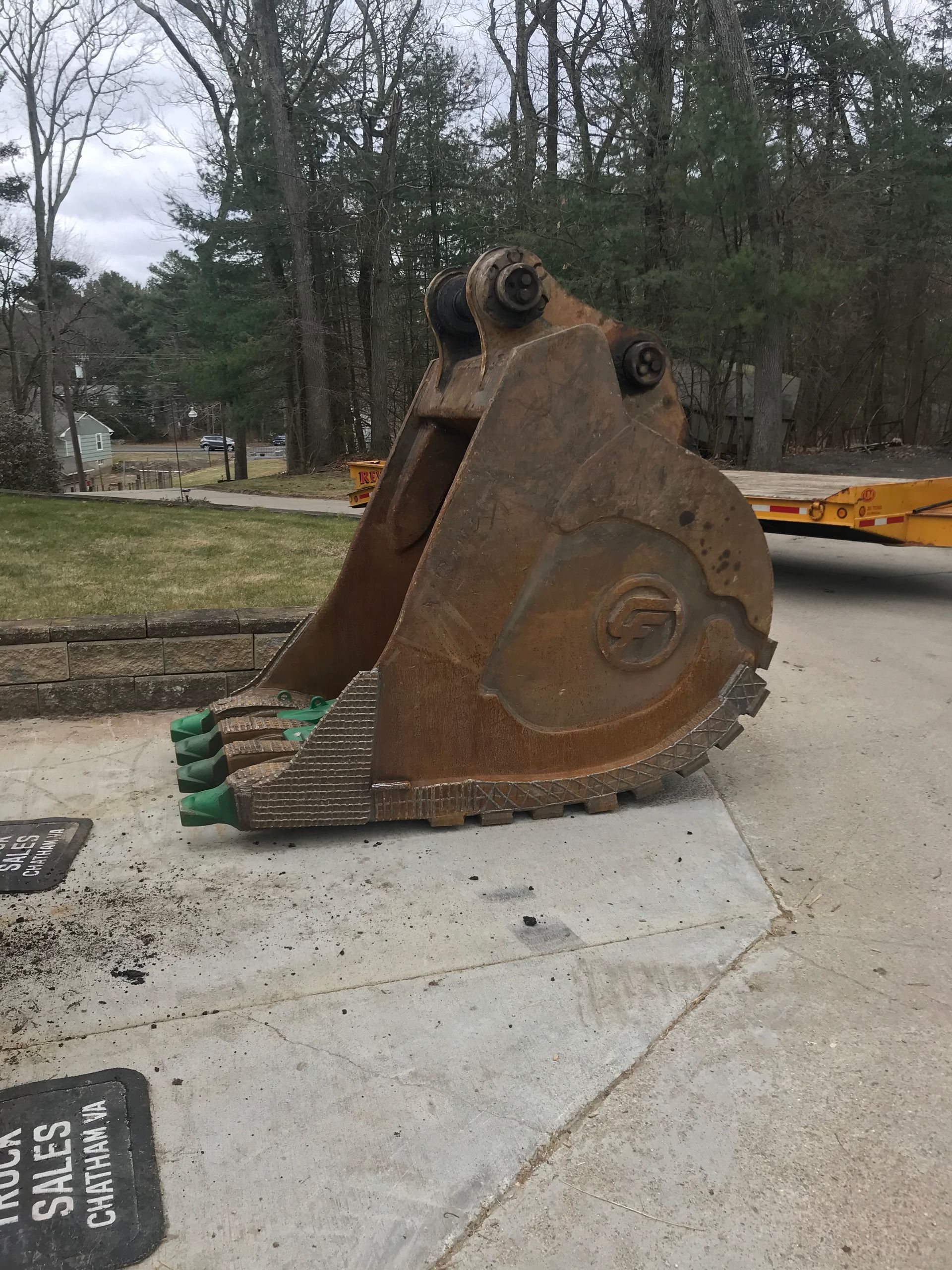 Large, rusted excavator bucket with green teeth sitting on a paved surface outdoors.