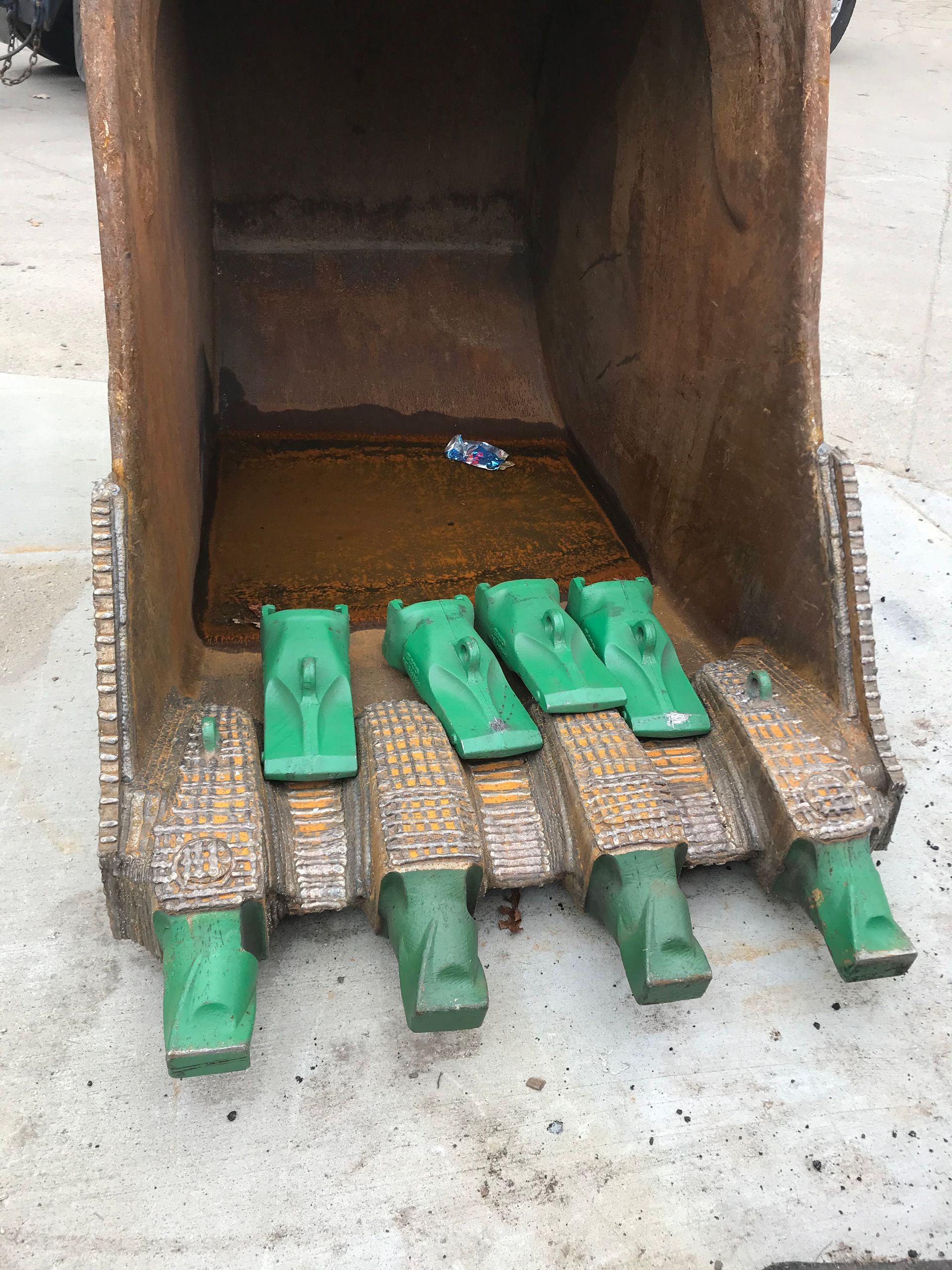 Close-up of a heavy excavator bucket with green metal teeth resting on a concrete surface.