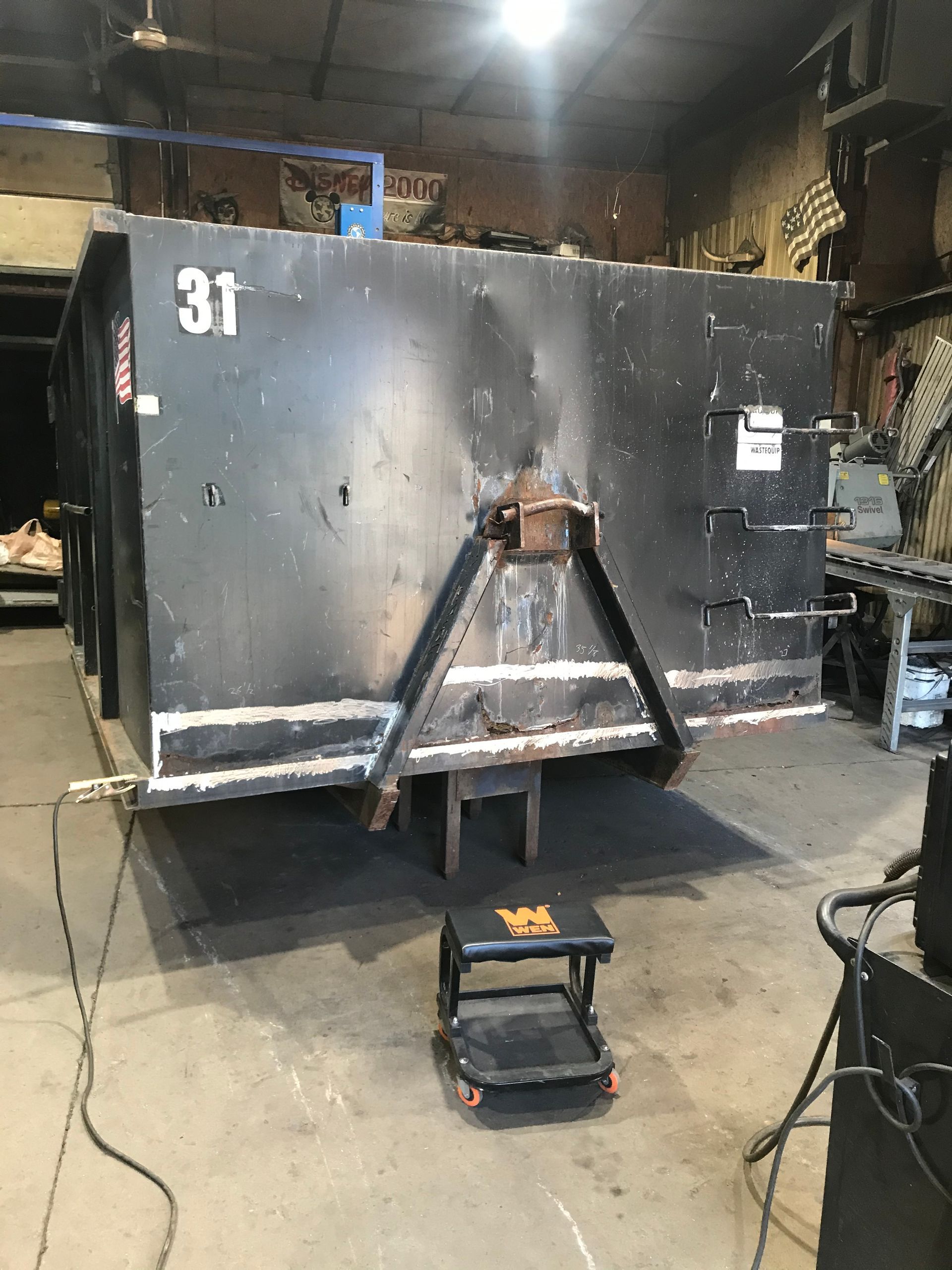 A black industrial dumpster with the number 31 sits in a workshop, with a small rolling step stool in front of it.