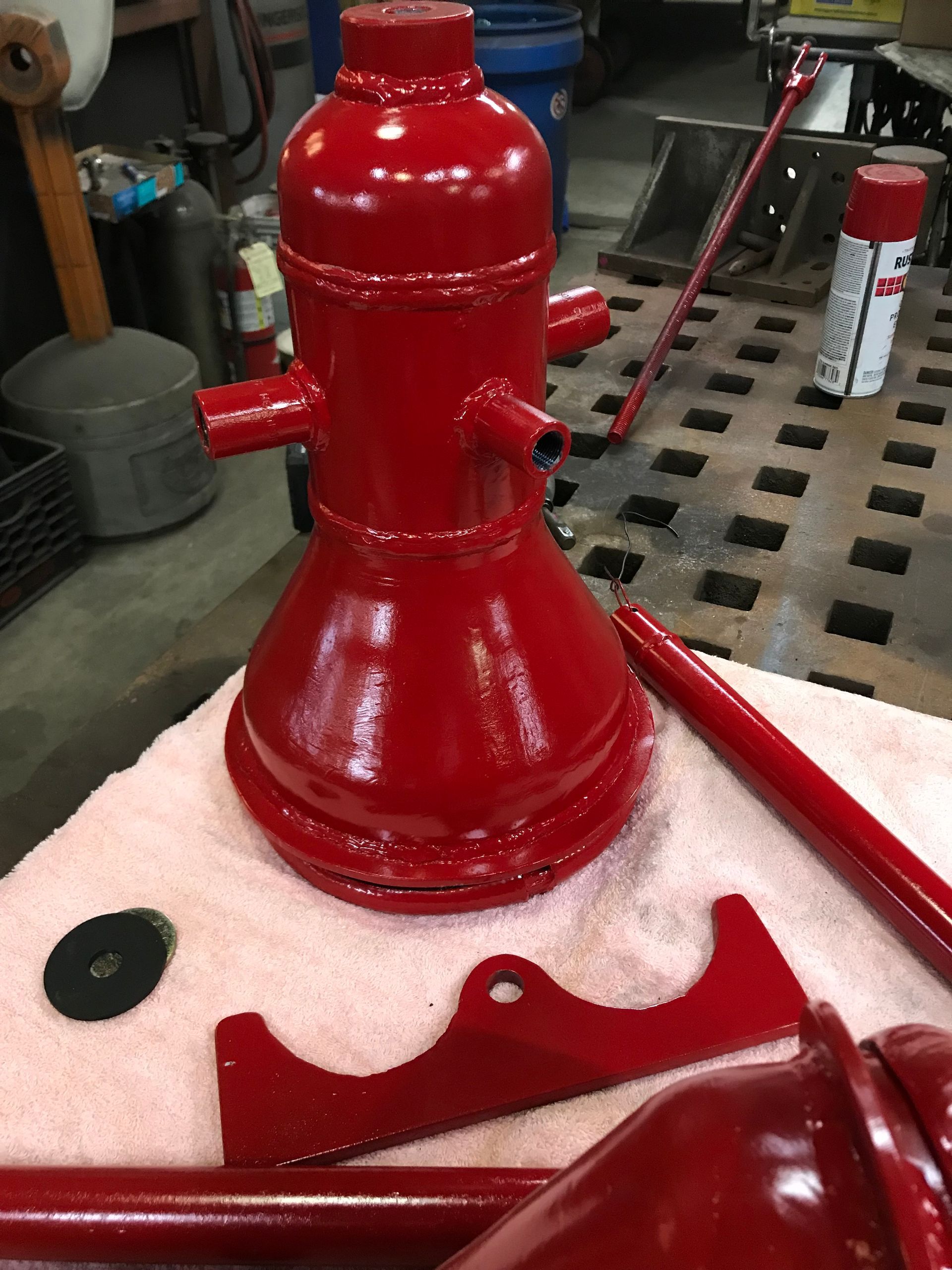 A newly painted, bright red vintage hydraulic jack stands on a pink towel alongside its disassembled metal parts.