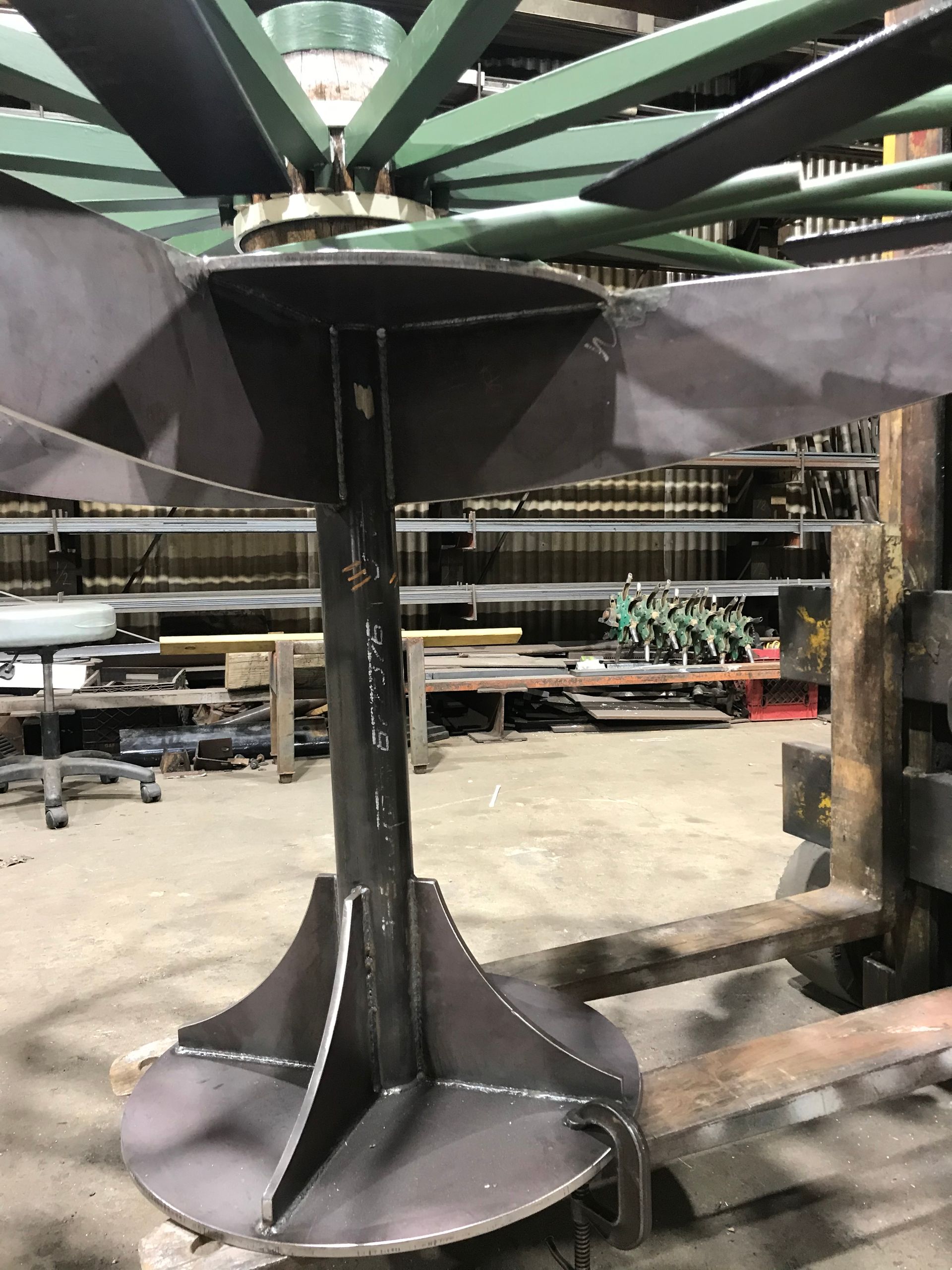 A metal stand with a round base and gussets, topped by a circular plate supporting green fan-like blades in a workshop.