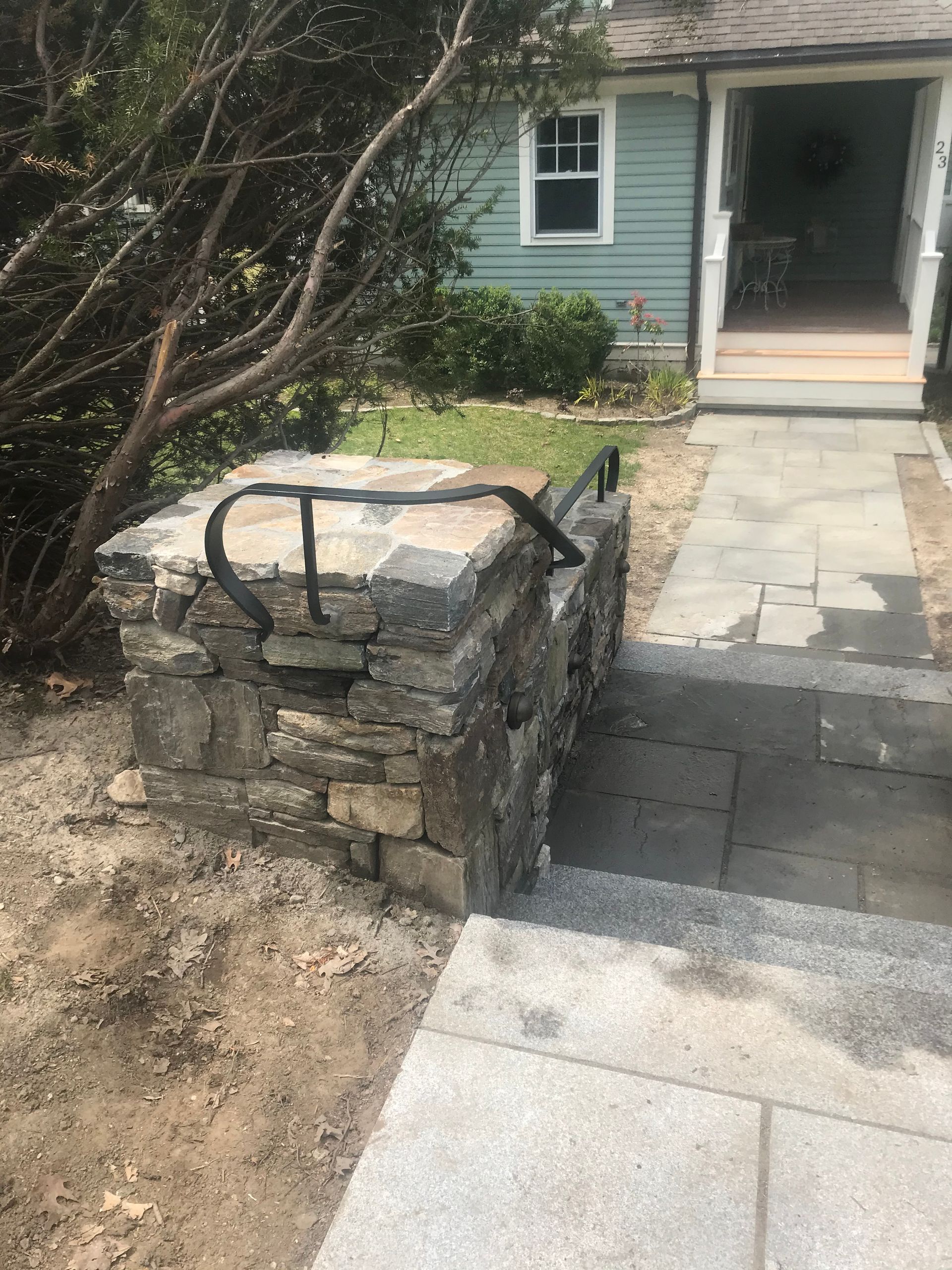 Stone stairs with a black metal handrail leading to a house entrance with light green siding and a stone walkway.
