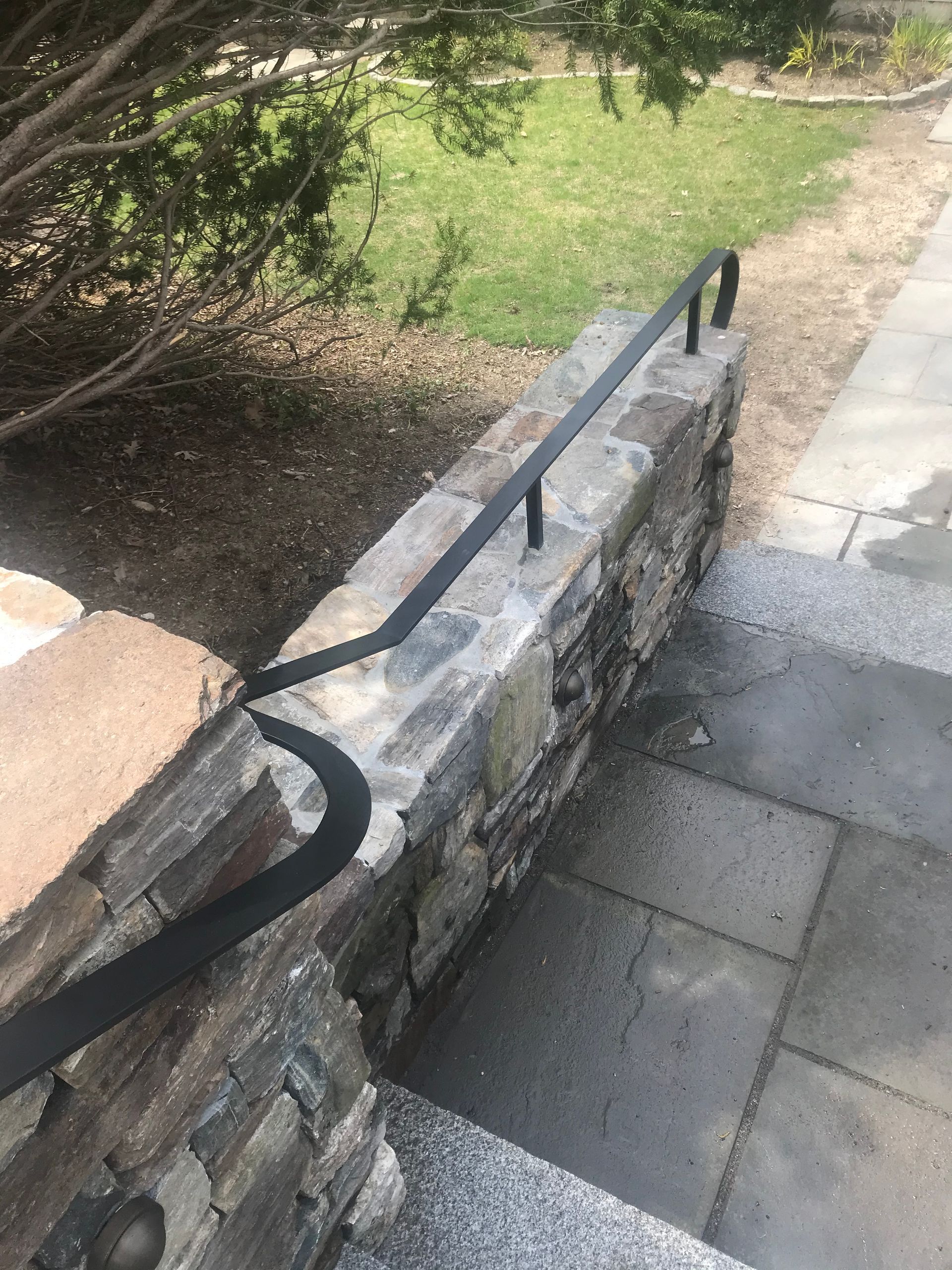 A black metal handrail mounted to a stone retaining wall next to outdoor stone steps.