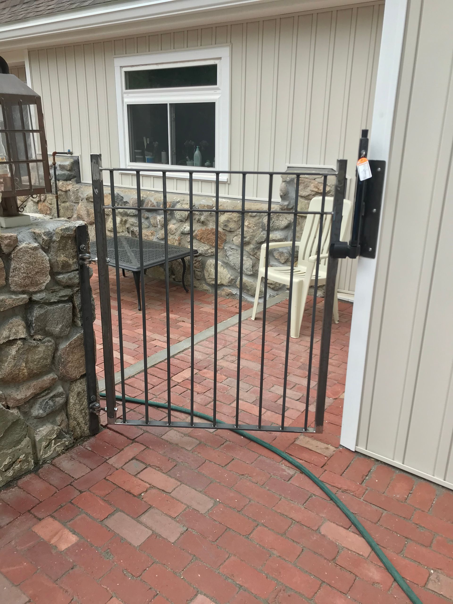 A black metal gate stands slightly ajar on a red brick patio in front of a stone wall and a white building.