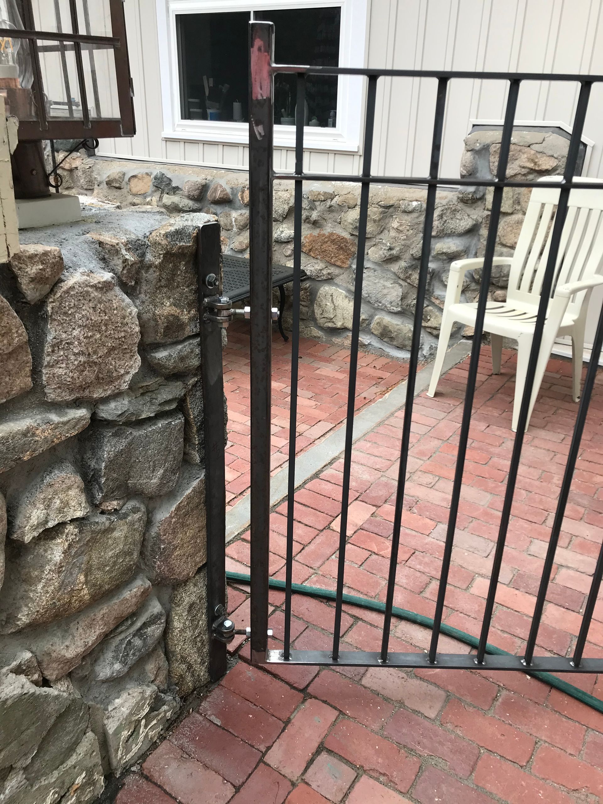 A black metal gate attached to a stone wall on a brick patio, with a white chair visible in the background.