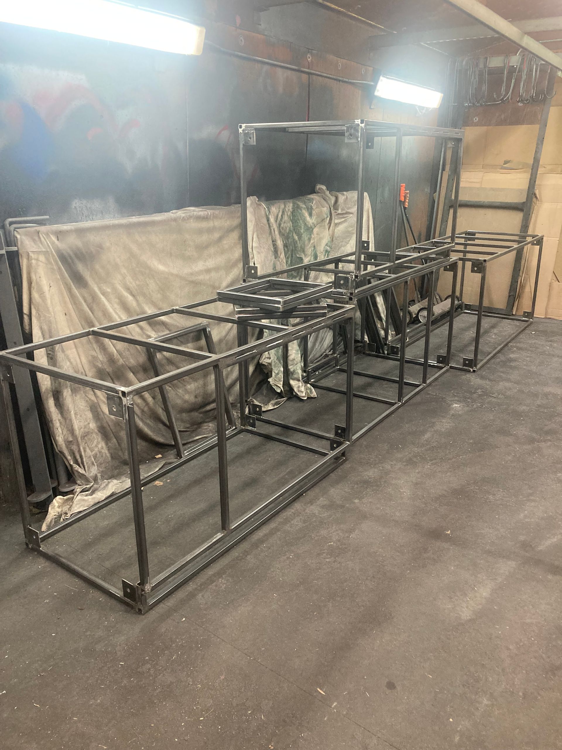 A rectangular metal frame structure made of dark rebar sits on a grey workshop floor against a draped, stained backdrop.