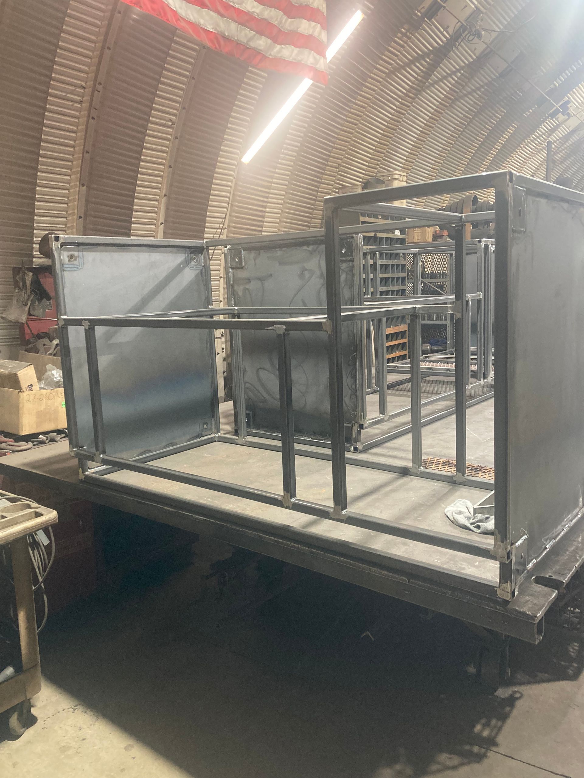 A metal frame for a custom vehicle bed or utility trailer sits on a workbench inside a metal workshop.