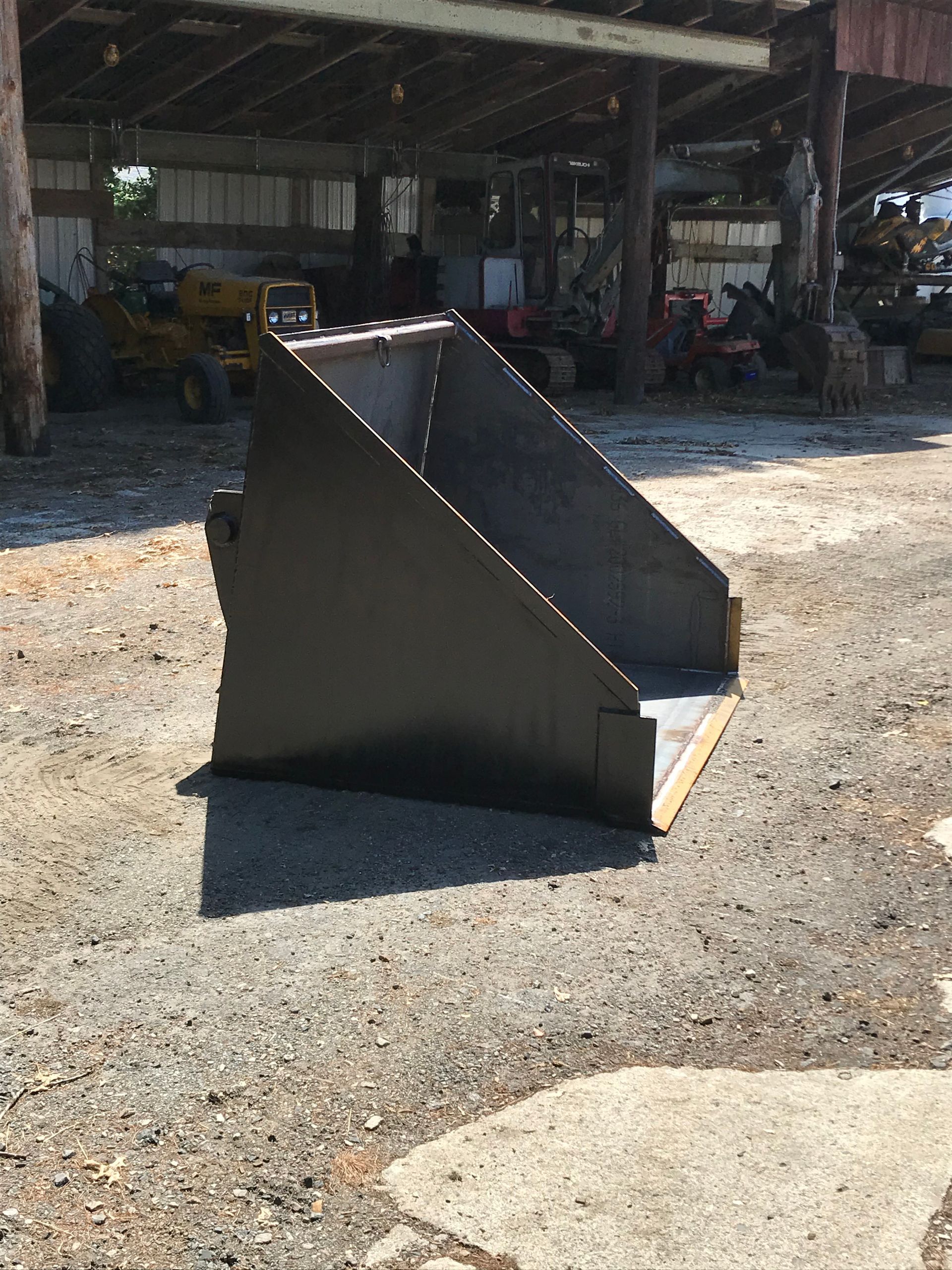 A black metal tractor loader bucket sits on a gravel lot in front of a rustic wooden shed.