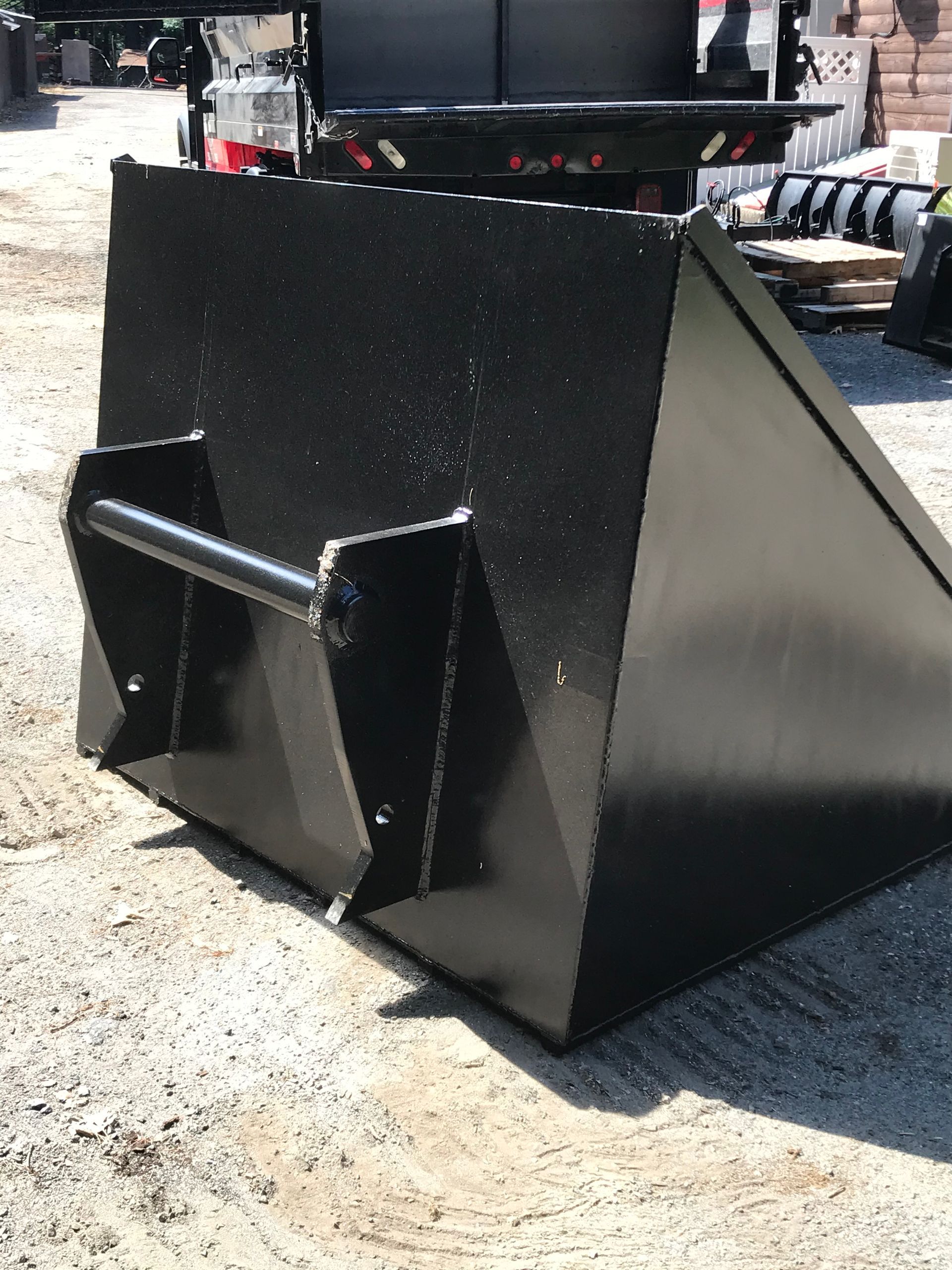 A glossy black metal loader bucket for a tractor or skid steer sits on a gravel surface.