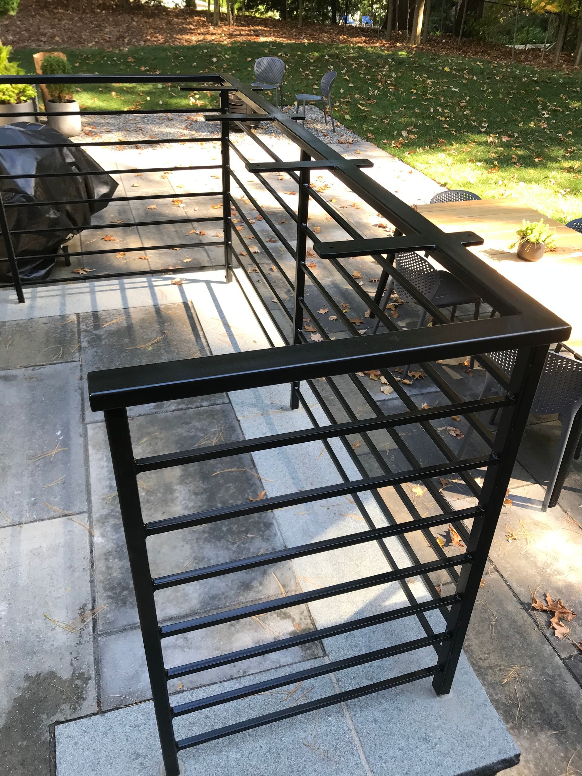 Black metal railing with horizontal bars installed on a light-colored stone patio, overlooking a grassy backyard.