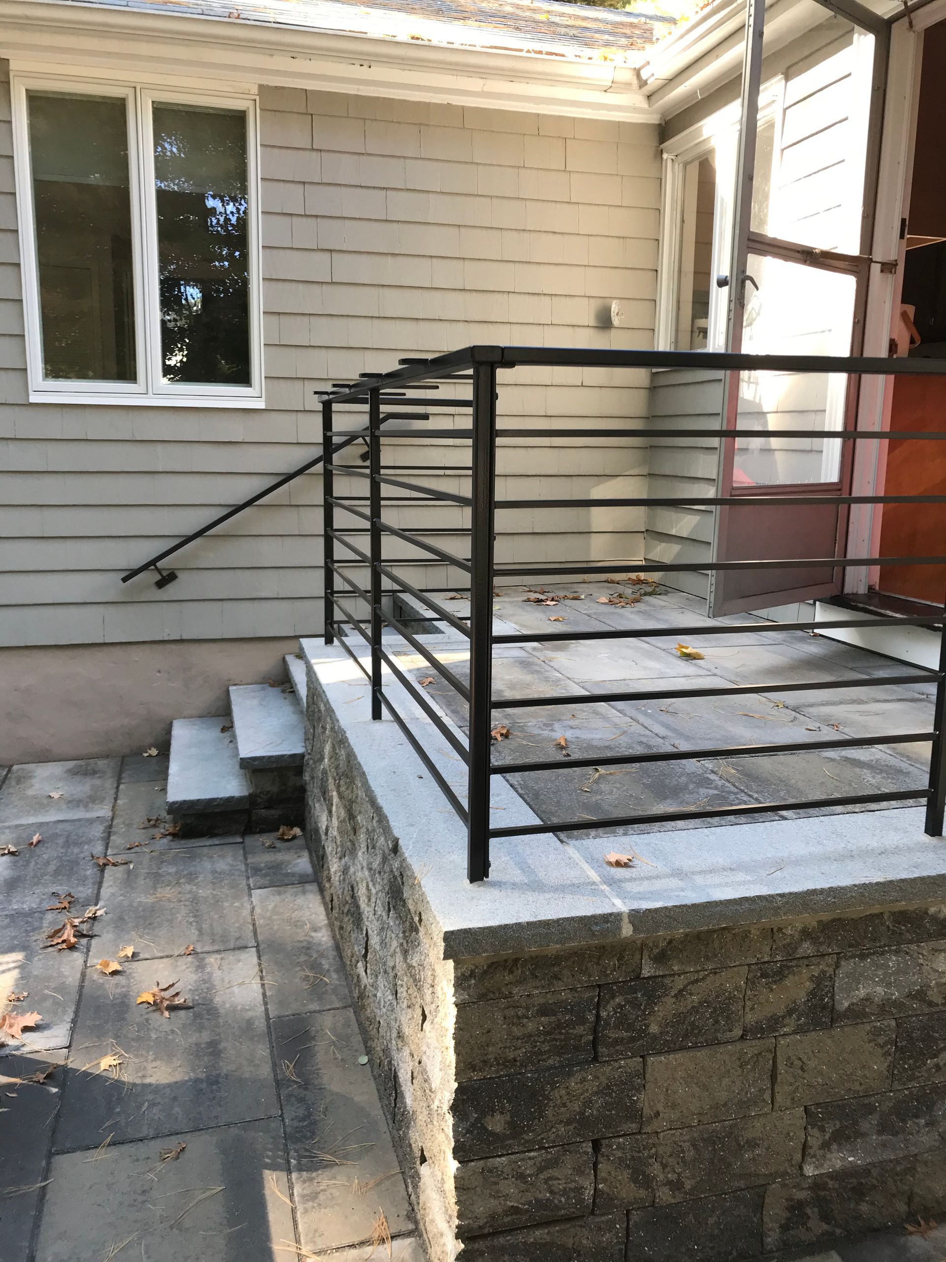 Black metal railing installed on a raised stone patio next to a house with concrete steps.