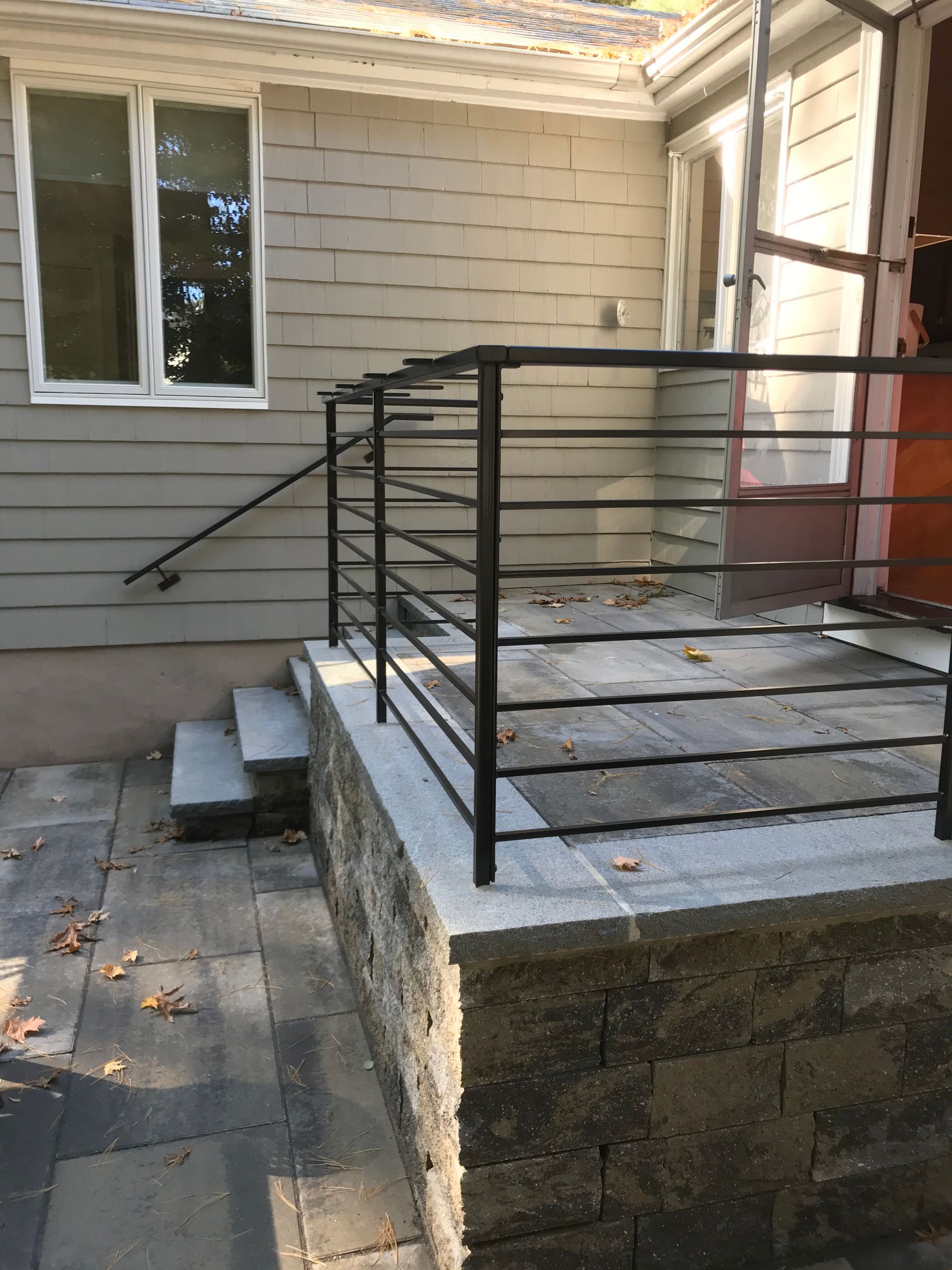 An outdoor stone patio with two concrete steps, a grey stone retaining wall, and a modern black metal railing.