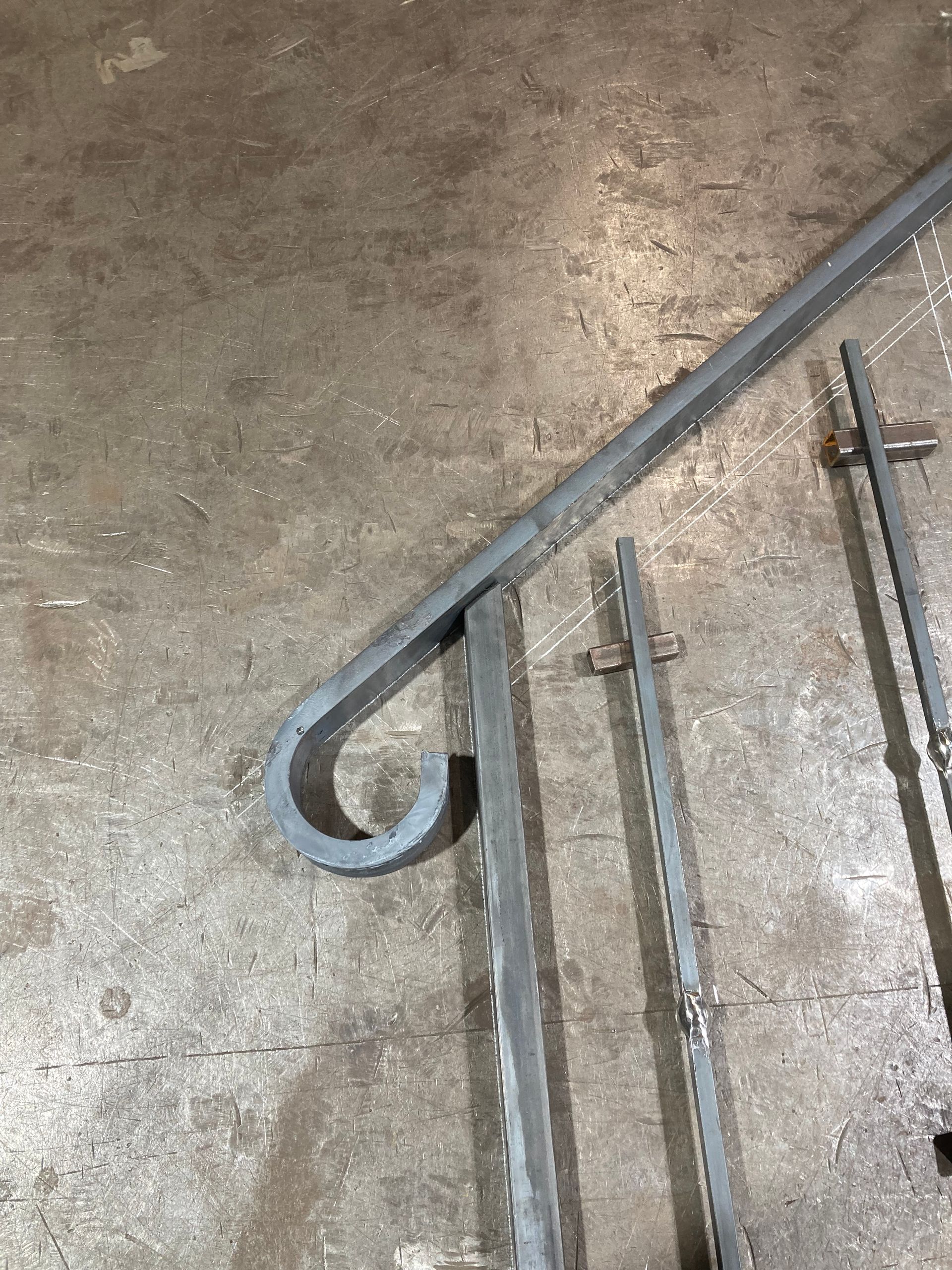 A gray, unfinished metal handrail with a scrolled end, positioned on a workshop floor next to vertical metal balusters.