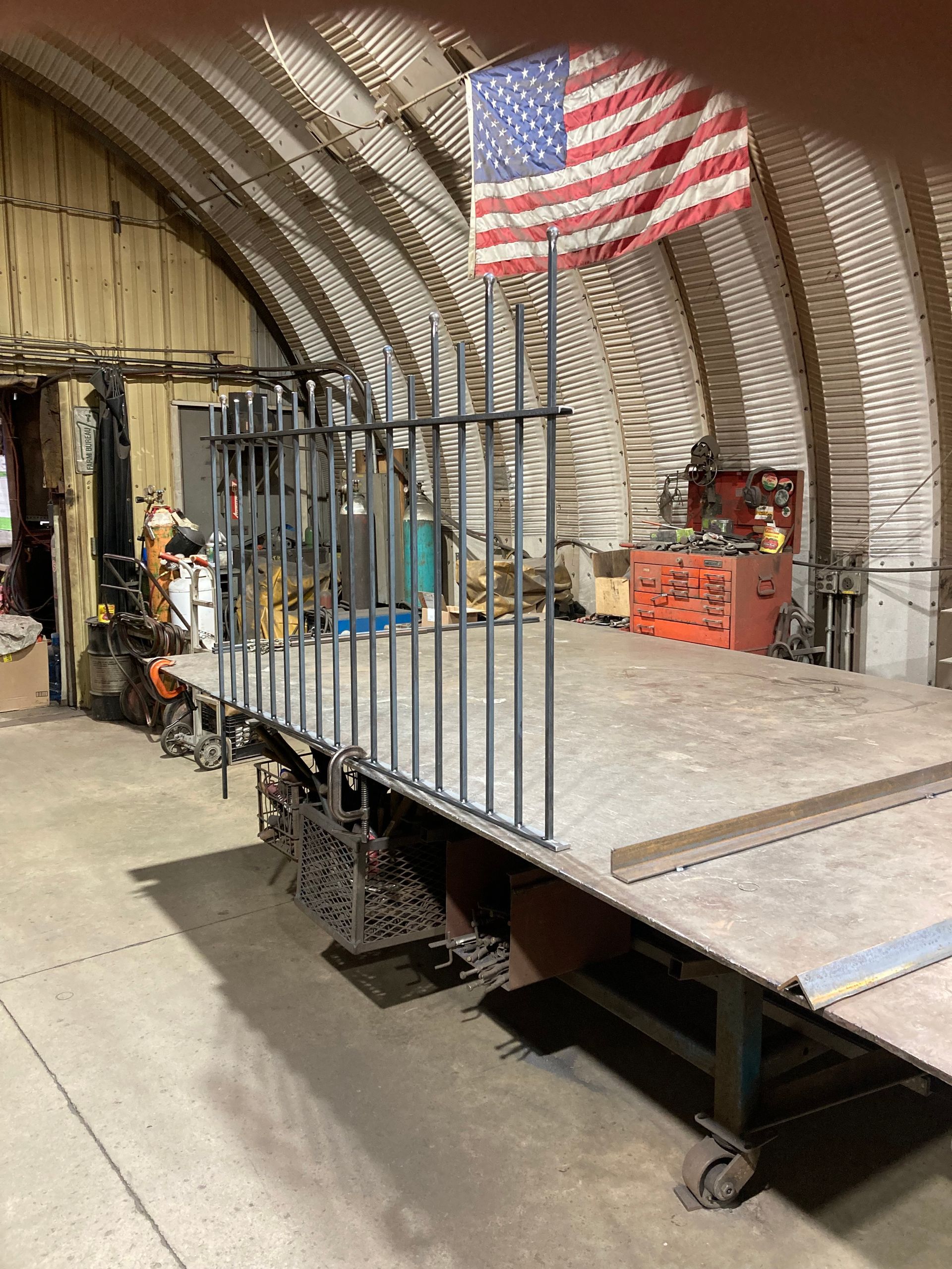 A metal fence section sits on a large, wheeled industrial workbench inside a workshop with a US flag on the corrugated wall.
