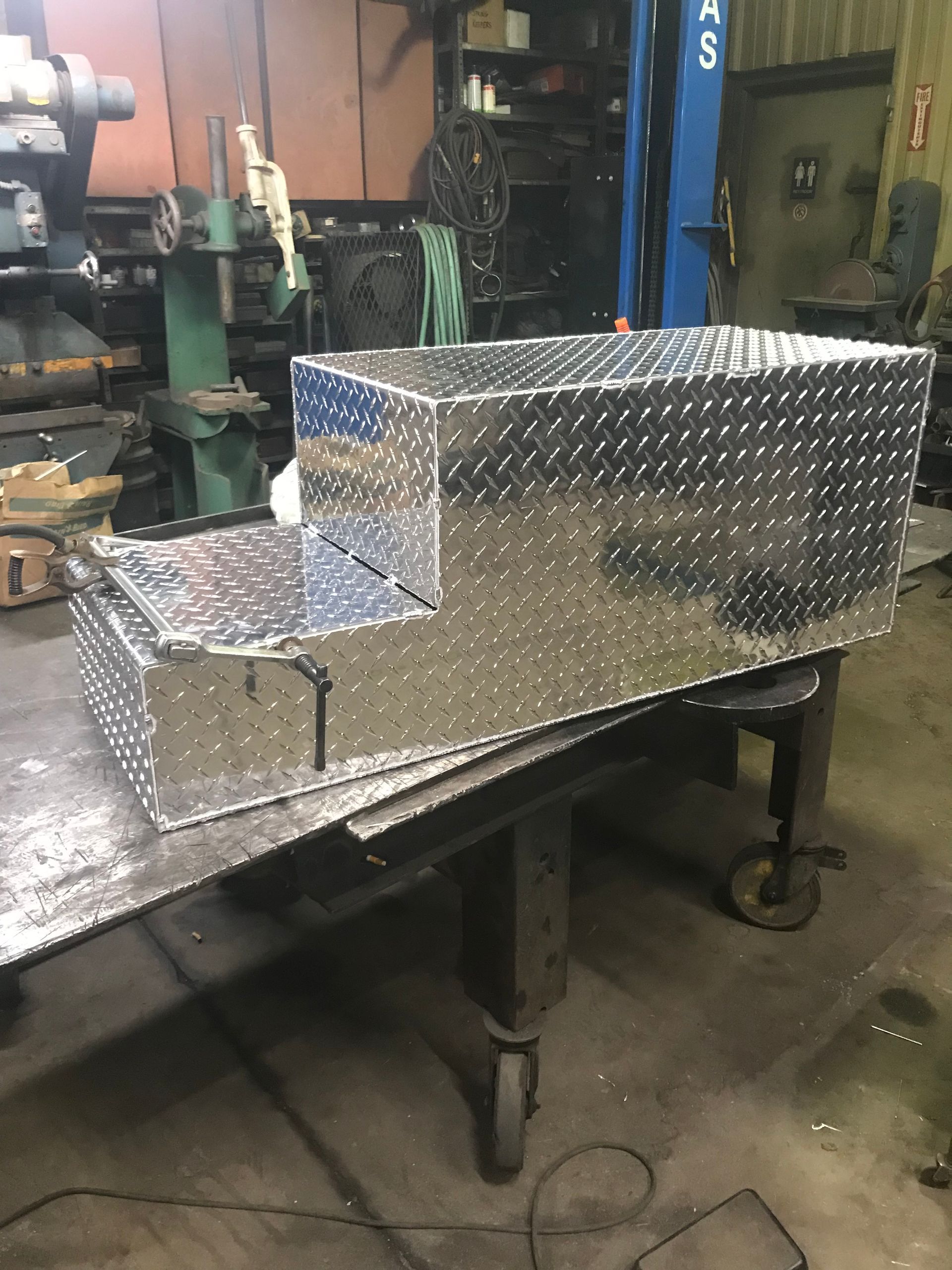 A diamond plate aluminum truck toolbox with a tiered, stepped design, sitting on a work bench in a shop.