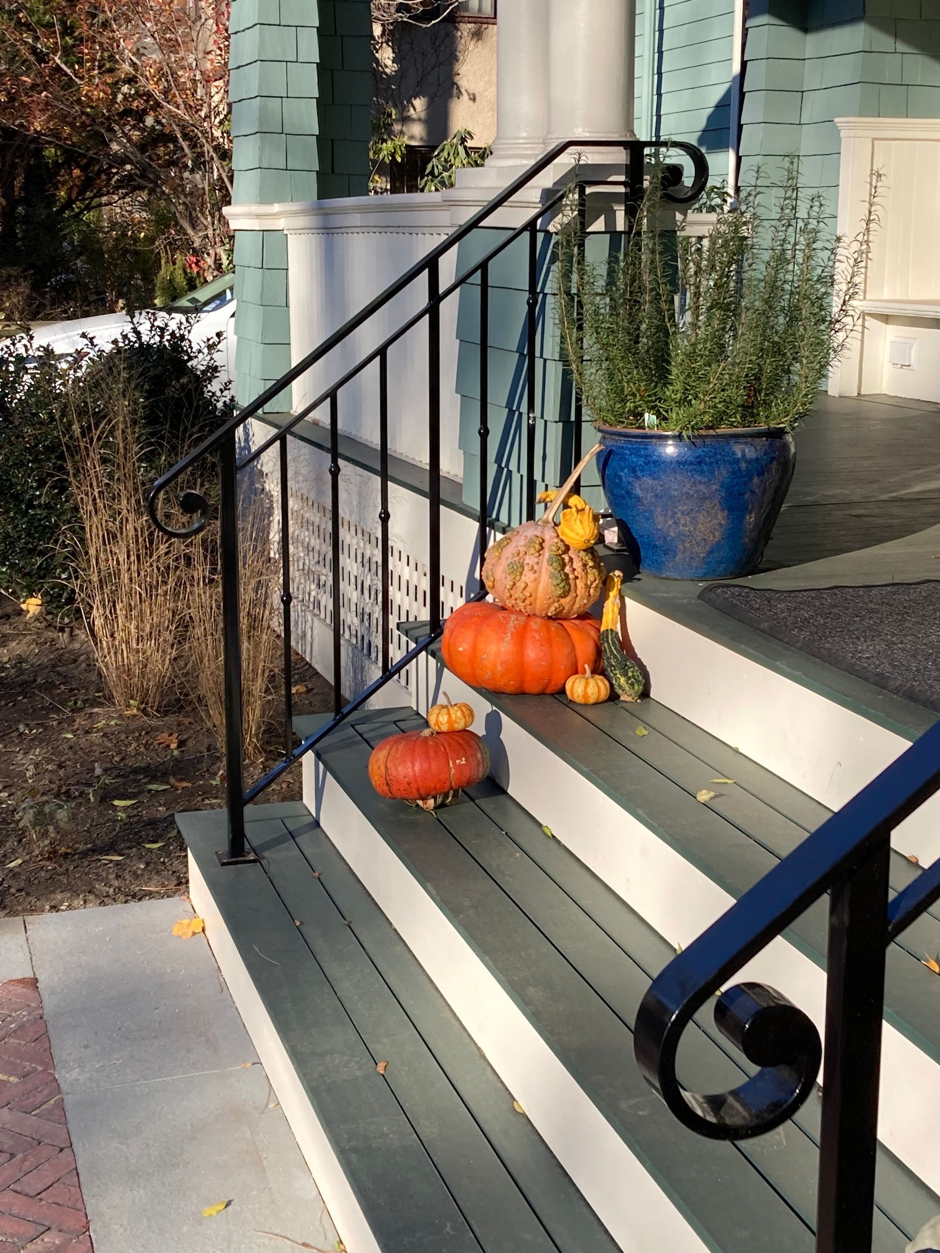 Orange pumpkins sit on the steps of a teal porch next to a black iron railing and a blue planter with green foliage.