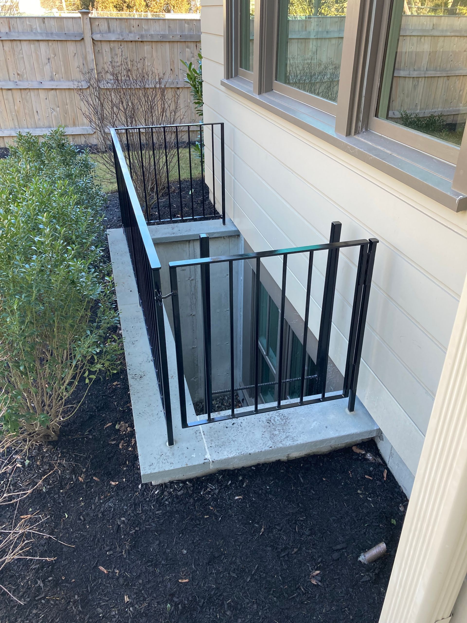 A below-grade basement window well exterior with a concrete border and black metal safety railing, set against a house.