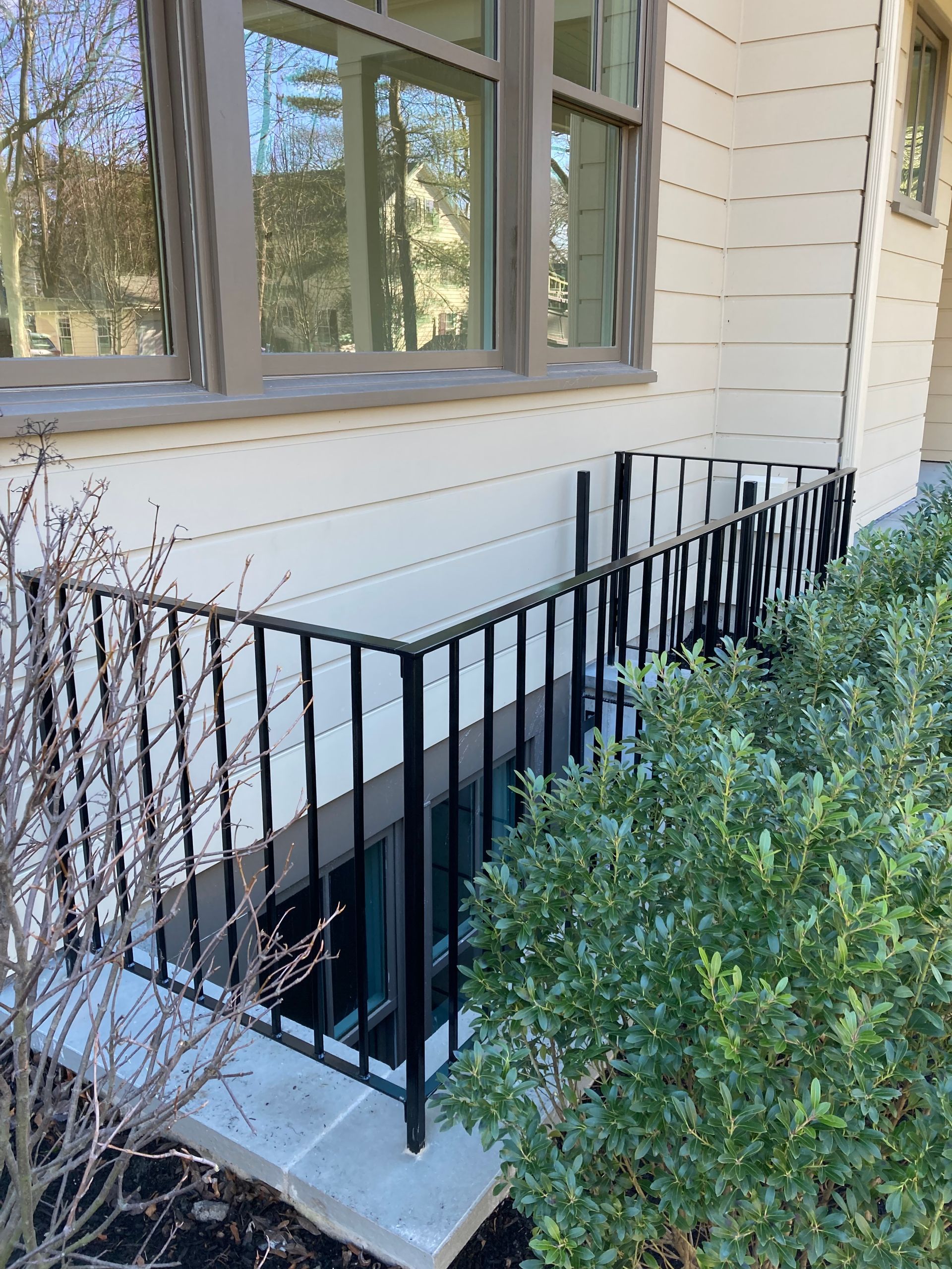 A black metal railing surrounds a basement window well on the side of a light-colored house near a green bush.