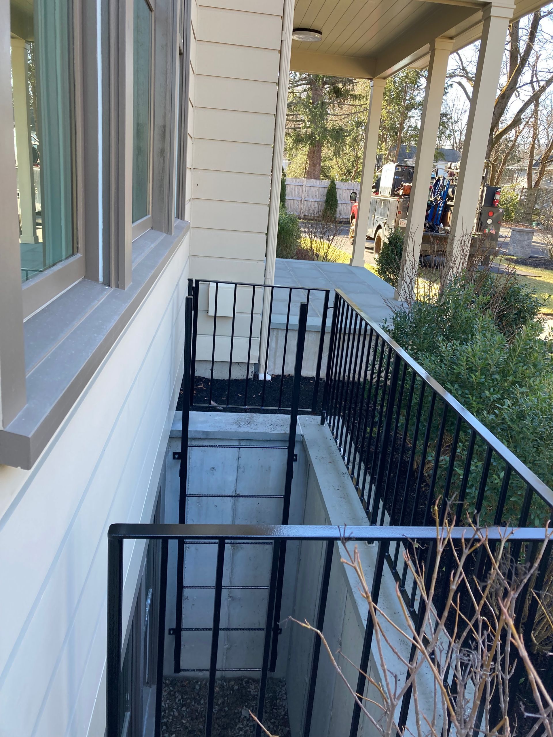 A basement egress window well with a black metal ladder and railing, set against the side of a light-colored house.
