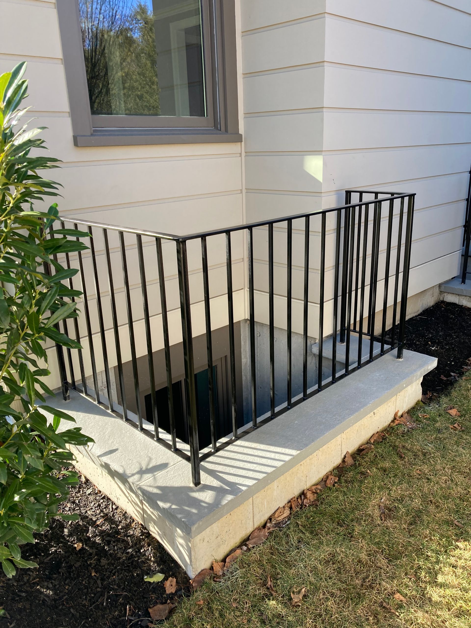 A black metal railing surrounds a concrete egress window well set against the exterior wall of a building.