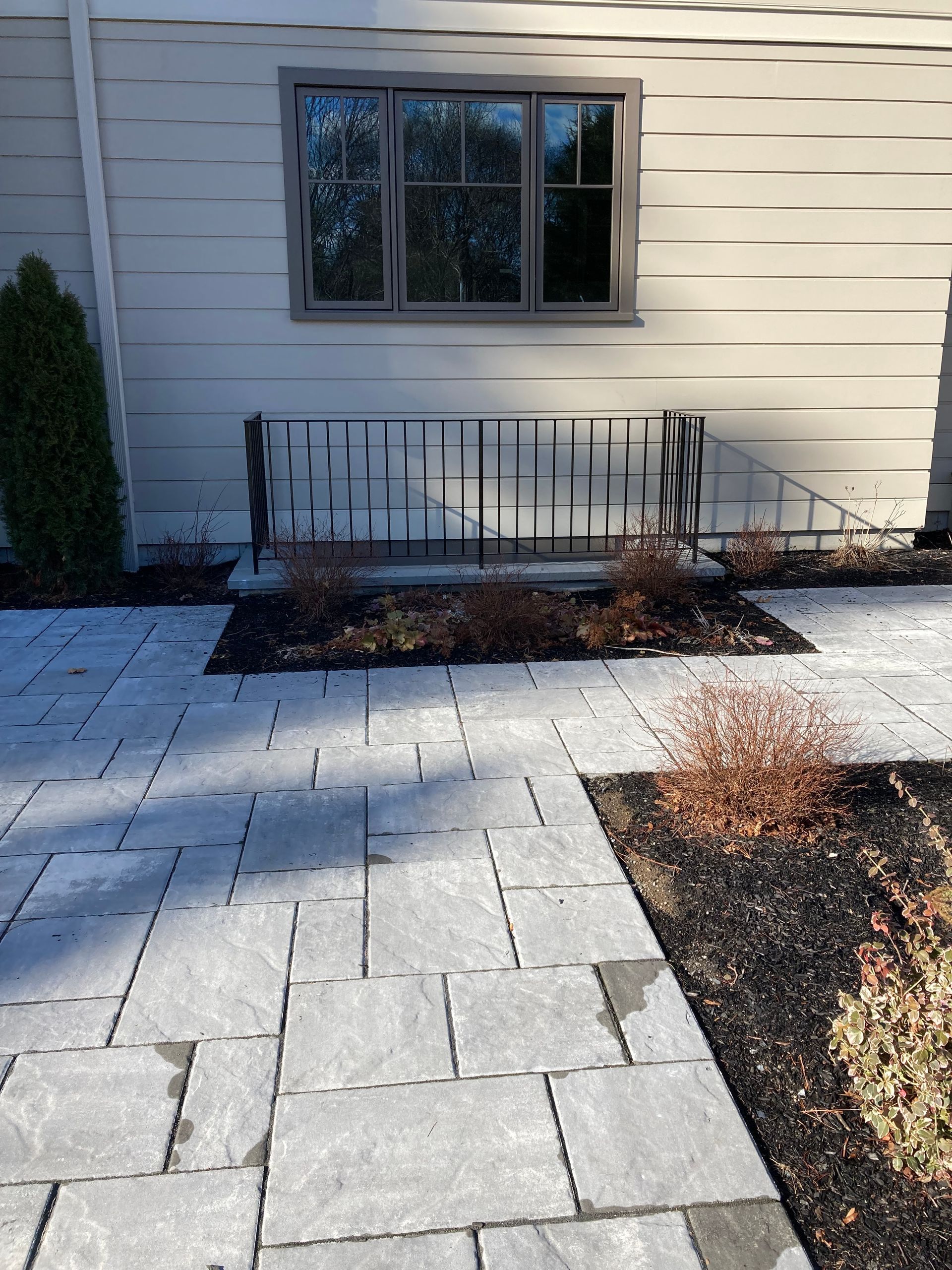 A gray stone patio leads to a light-colored building with a small metal railing and landscape beds with young shrubs.