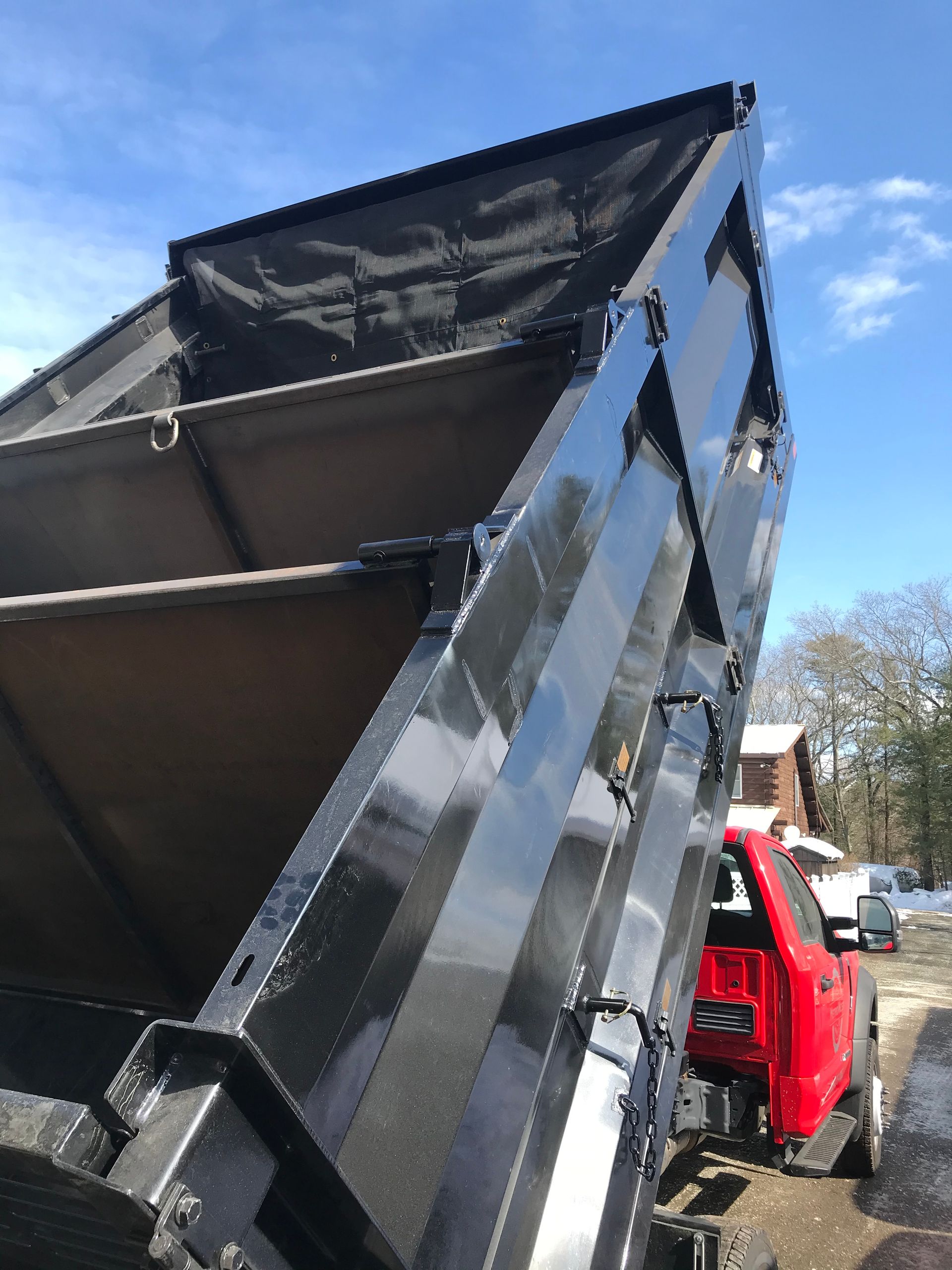 A red truck with its large, black metal dump bed raised high against a blue sky.