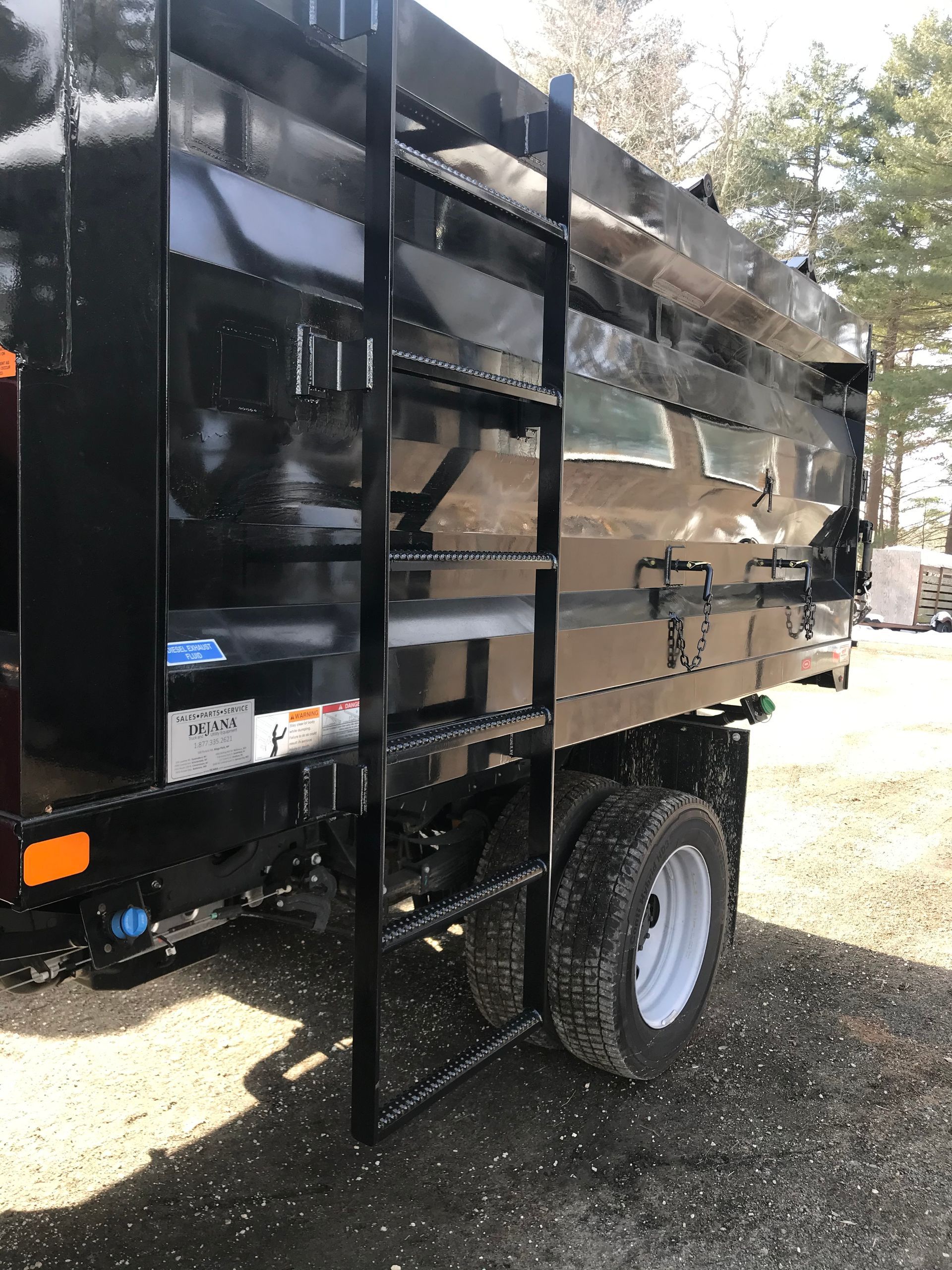 A black dump truck bed with an attached side ladder, parked on a gravel surface outdoors.