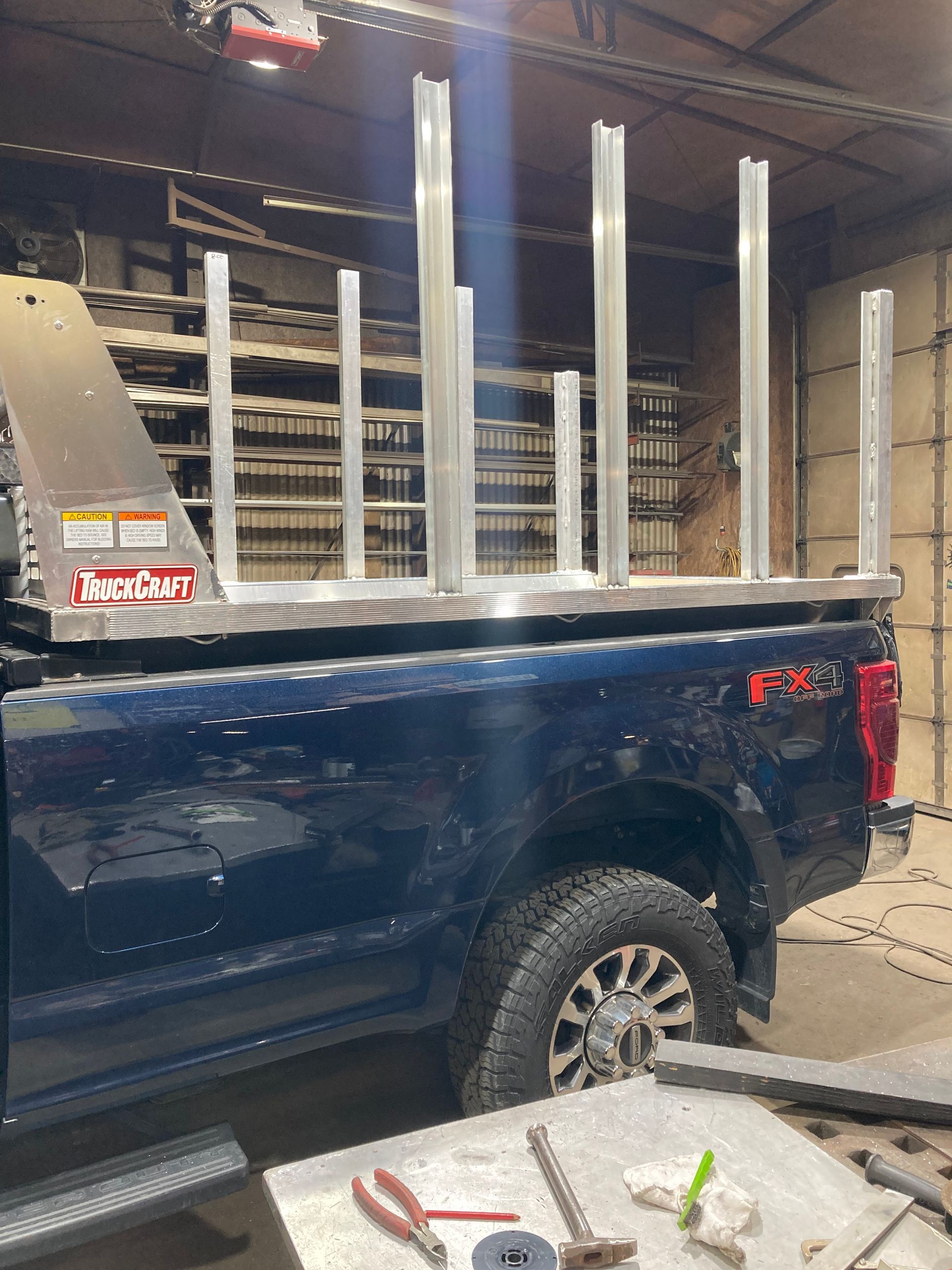 A blue pickup truck in a workshop features a newly installed aluminum lumber rack with upright posts on the truck bed.