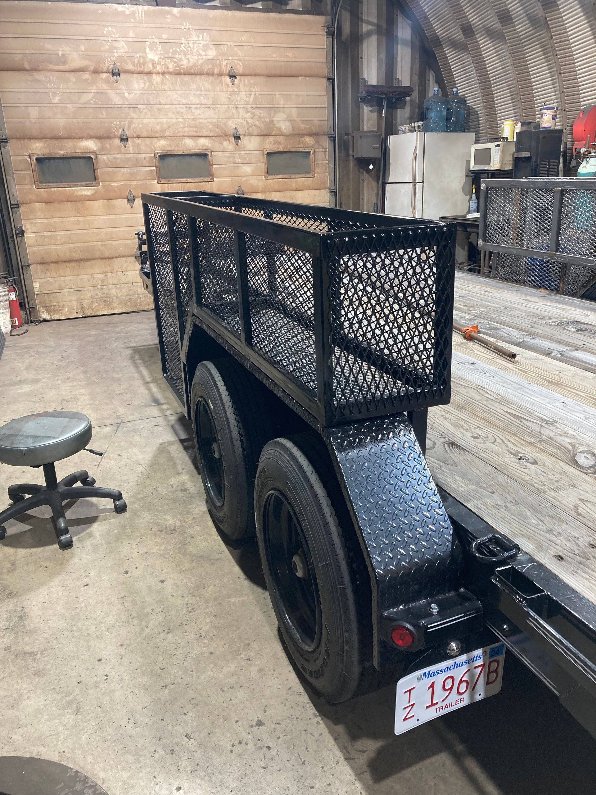 A black tandem-axle utility trailer with a mesh side wall and diamond-plate fenders inside a workshop.