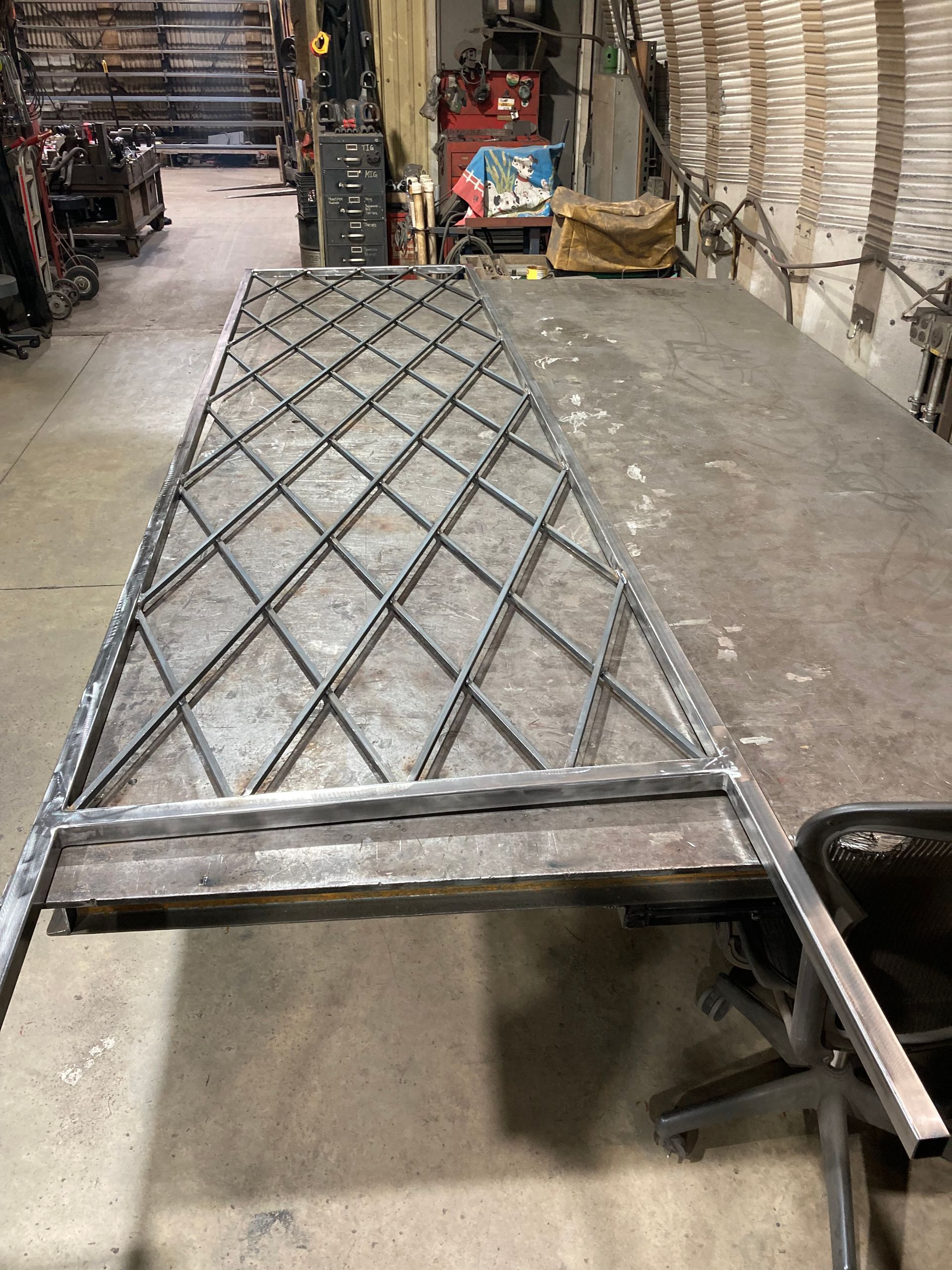 A metal frame with a diamond-patterned lattice grid sits on a workshop workbench, surrounded by metalworking equipment.