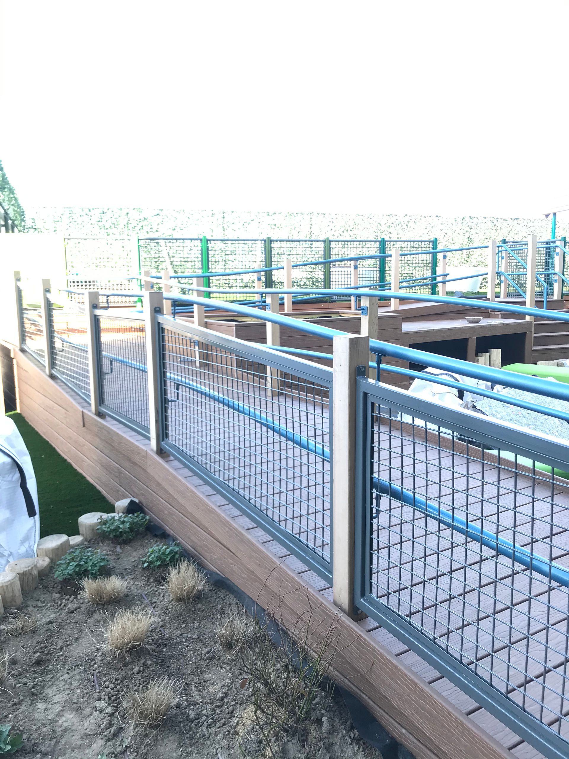 A multi-level outdoor wooden deck with metal railings and grid panels overlooks a landscaped area with small shrubs.
