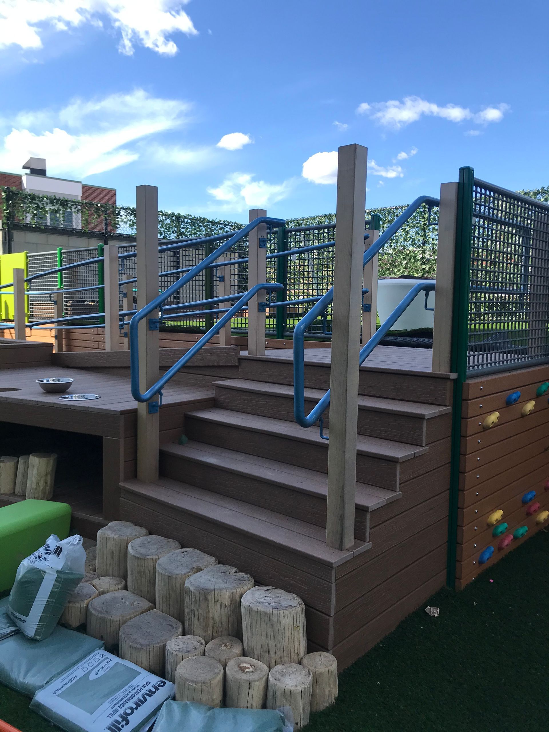 A brown wooden deck with stairs and blue railings, featuring a rock climbing wall on the side and a log step area outdoors.